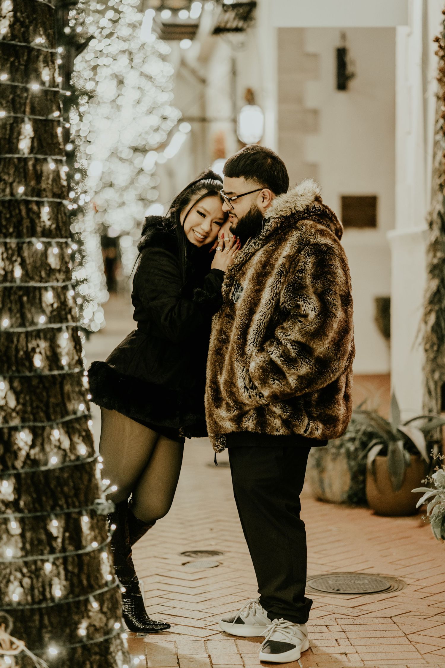 Couple sharing an intimate moment leaning against light-wrapped trees on a brick walkway at night.