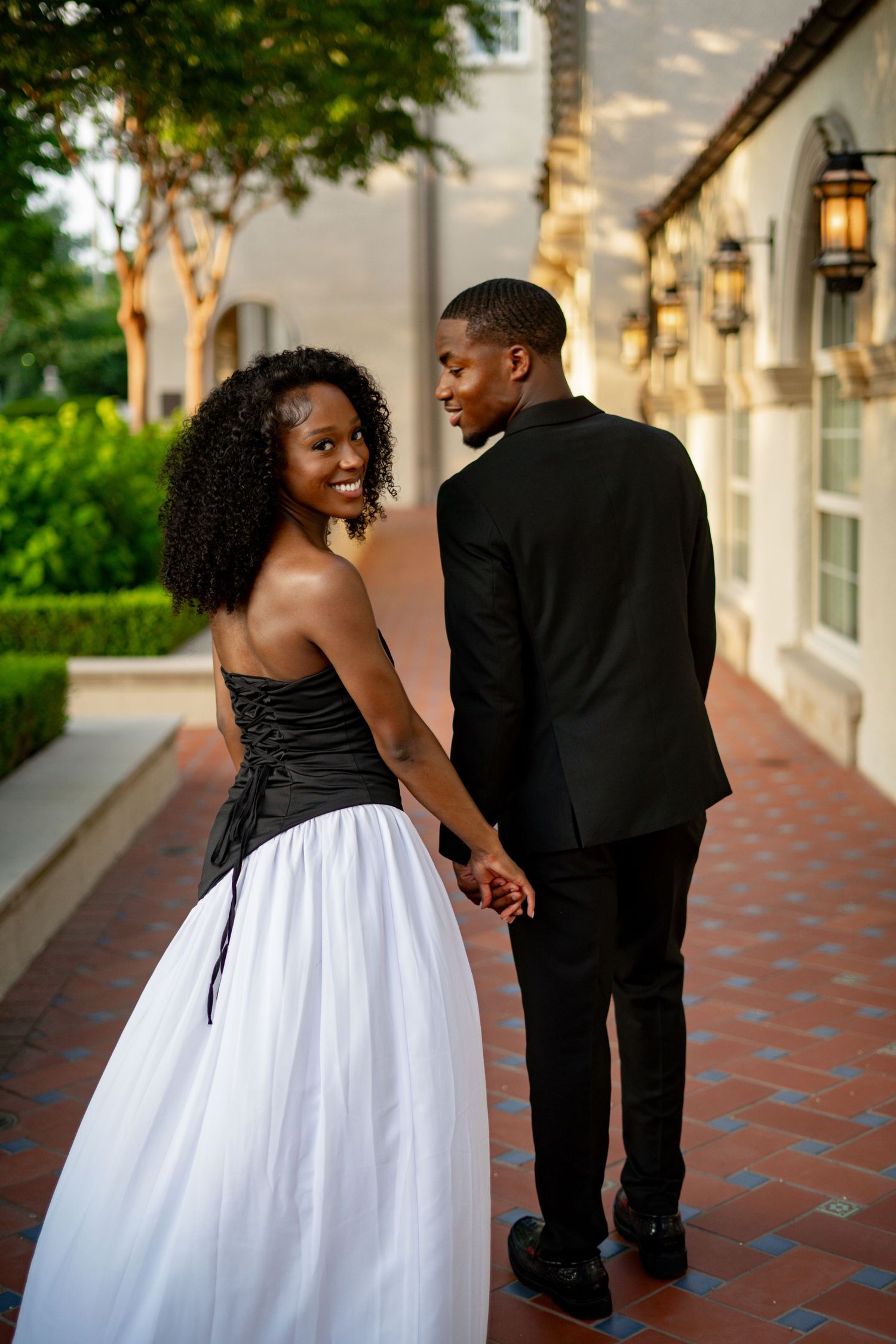 Smiling Black woman in black and white strapless gown holds hands with man in black suit in outdoor corridor.