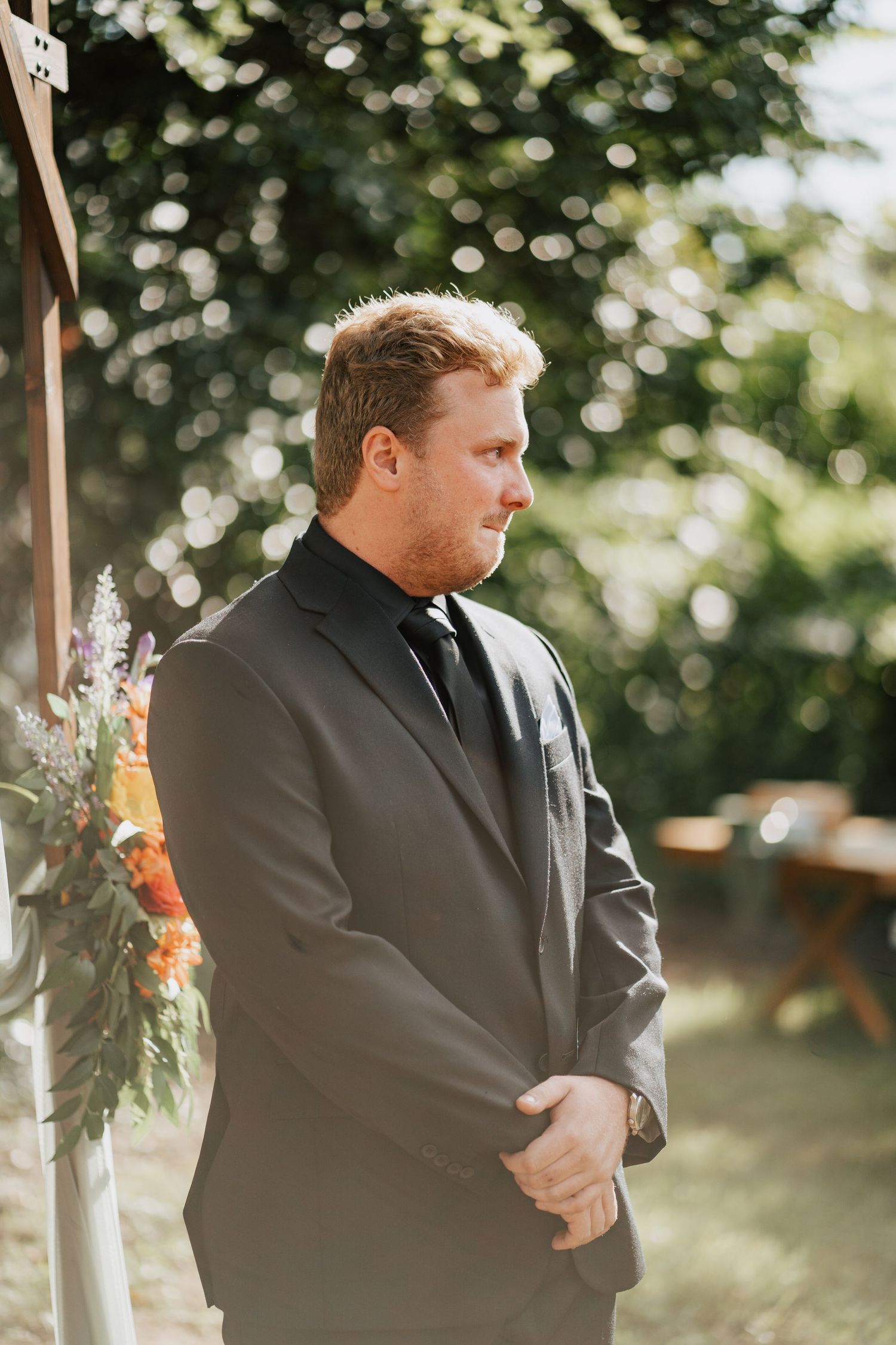 Groom in gray suit and black tie stands at outdoor wedding altar, looking emotional as he awaits his bride.