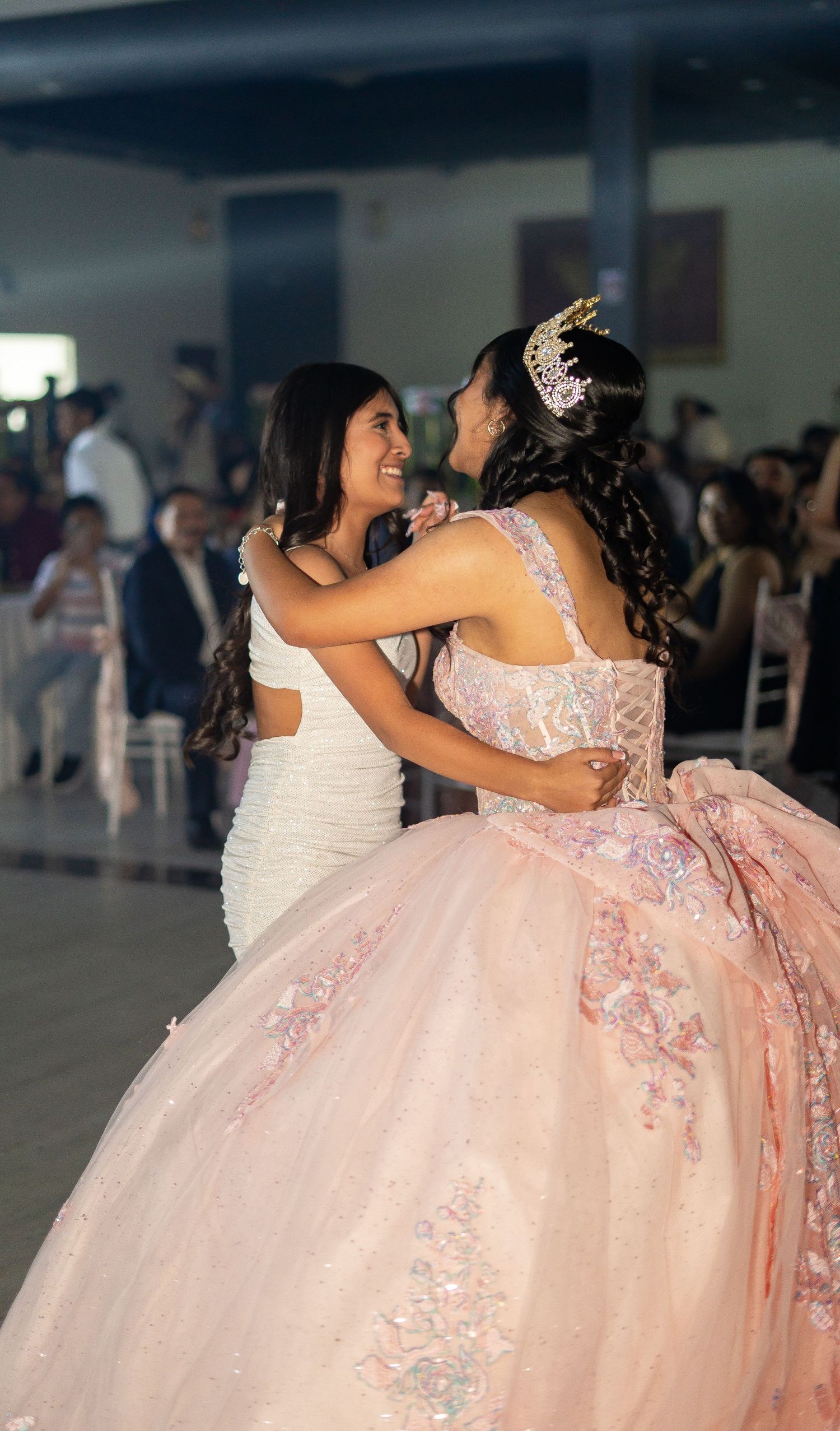 Two young women embrace and laugh during emotional quinceañera dance celebration at elegant venue.