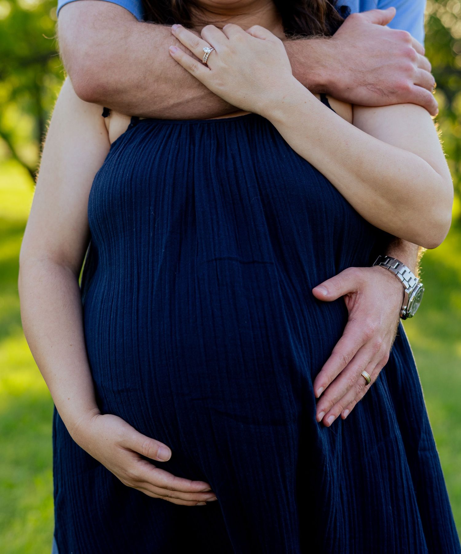 Close-up of pregnant woman in navy dress with partner embracing her from behind outdoors.