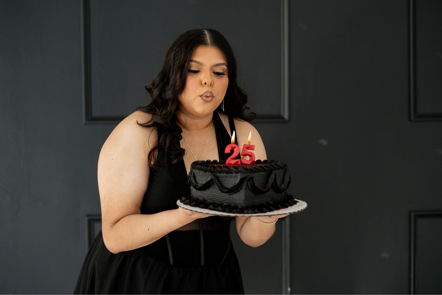 Woman in black dress blowing out number 25 candles on dark birthday cake.