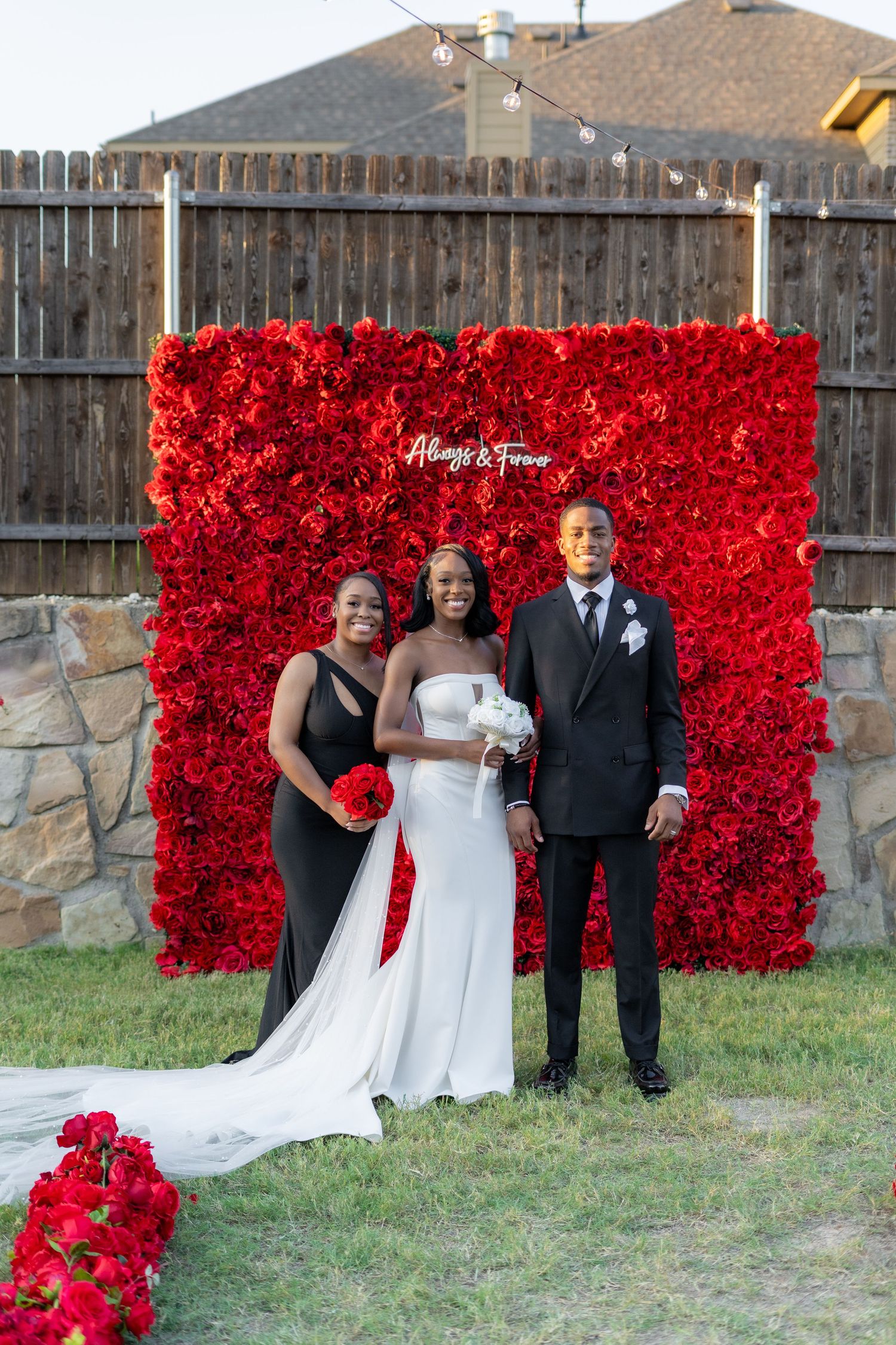 Bride groom and bridesmaid pose together in front of red rose flower wall at outdoor wedding.