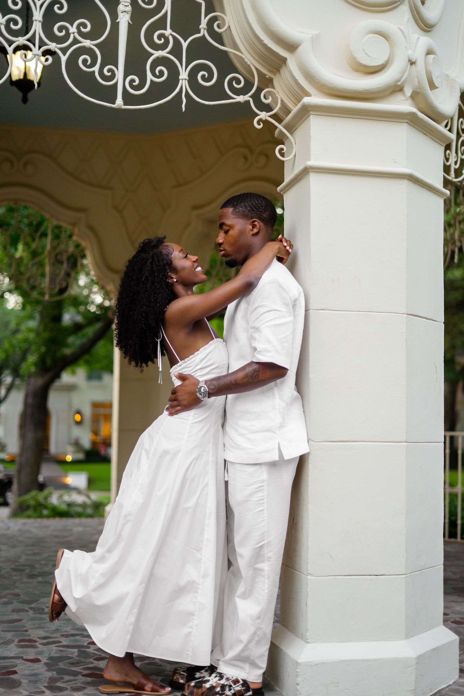 A Black couple in white wedding attire share a romantic embrace under an ornate white gazebo arch.