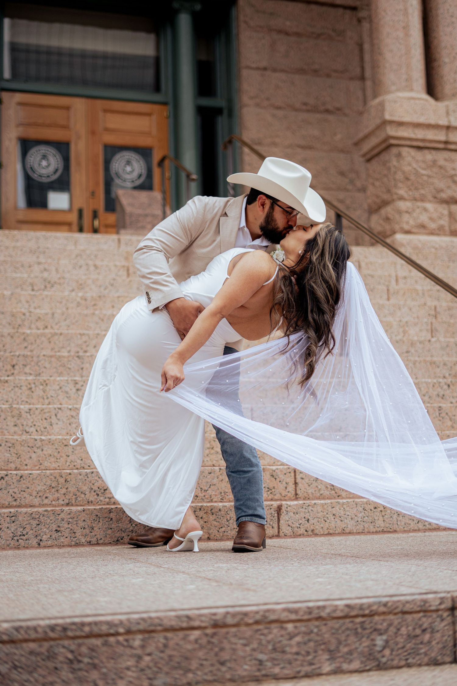 A groom in a white cowboy hat dips his bride in a white gown on stone steps outside a historic building.