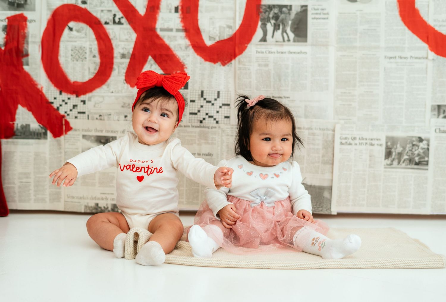 Two smiling babies in Valentine's Day outfits posed before a festive XOXO backdrop.