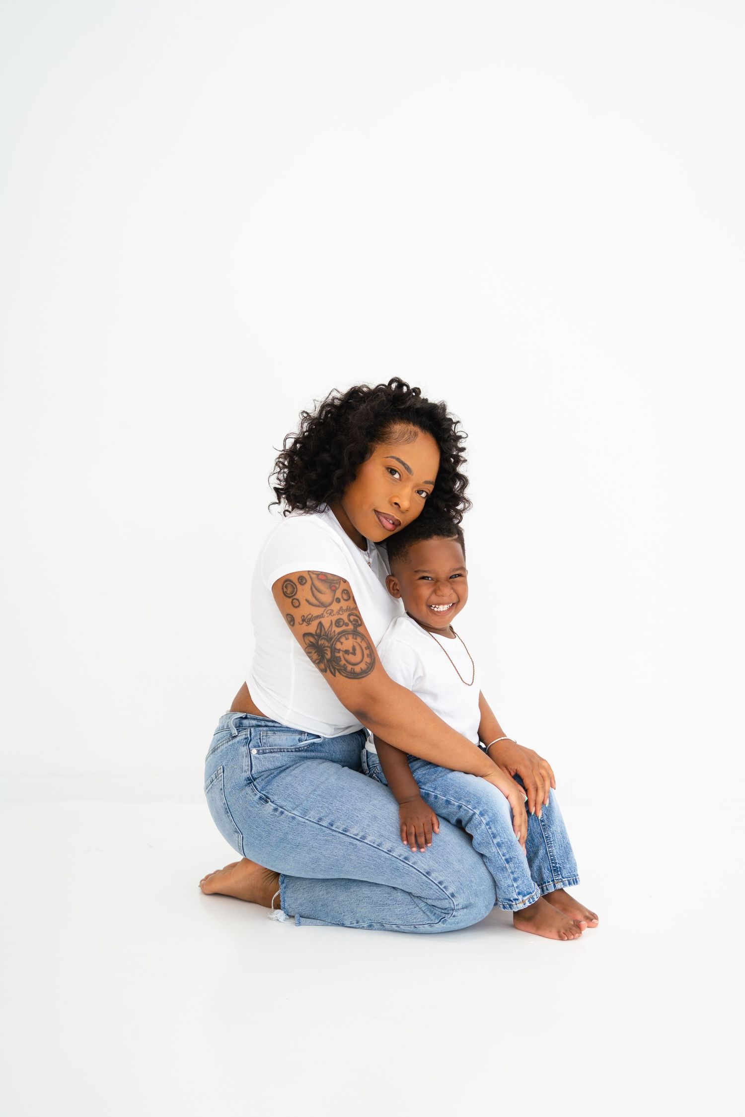 Mother and young son in matching white tees and jeans sharing a warm embrace on a white background.