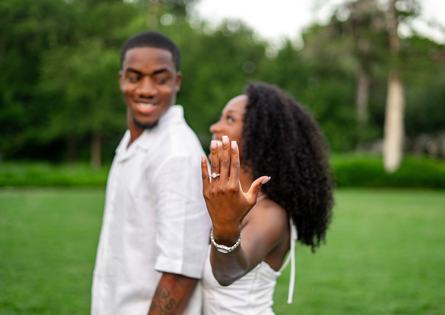 An engaged Black woman displays her engagement ring to the camera while her smiling partner stands behind her.