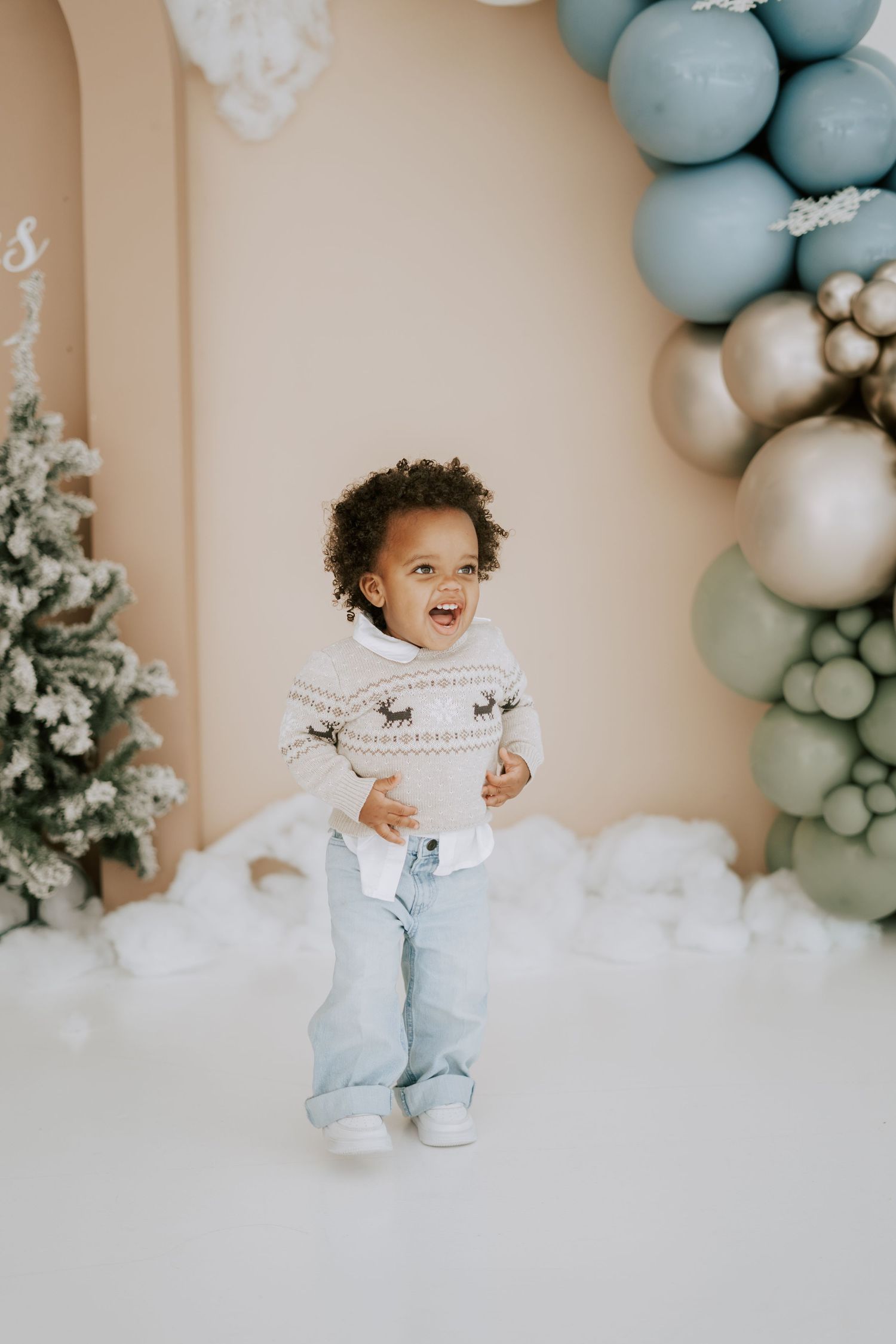 A happy toddler stands in front of a festive balloon arch with blue and gold Christmas decorations.