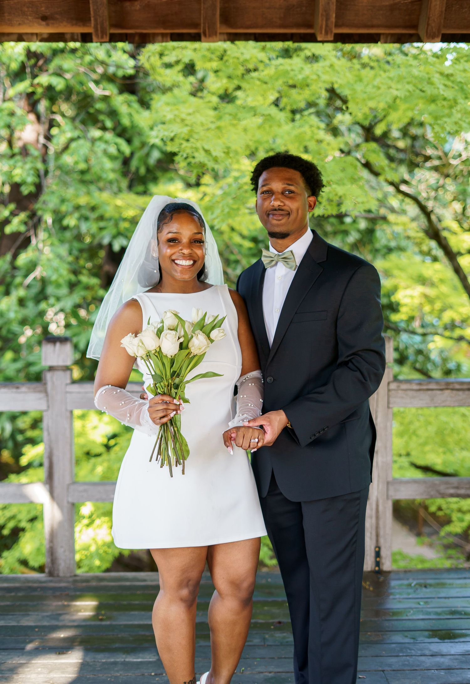 Bride in short white dress and veil holds tulip bouquet beside groom in black tuxedo at outdoor wedding.