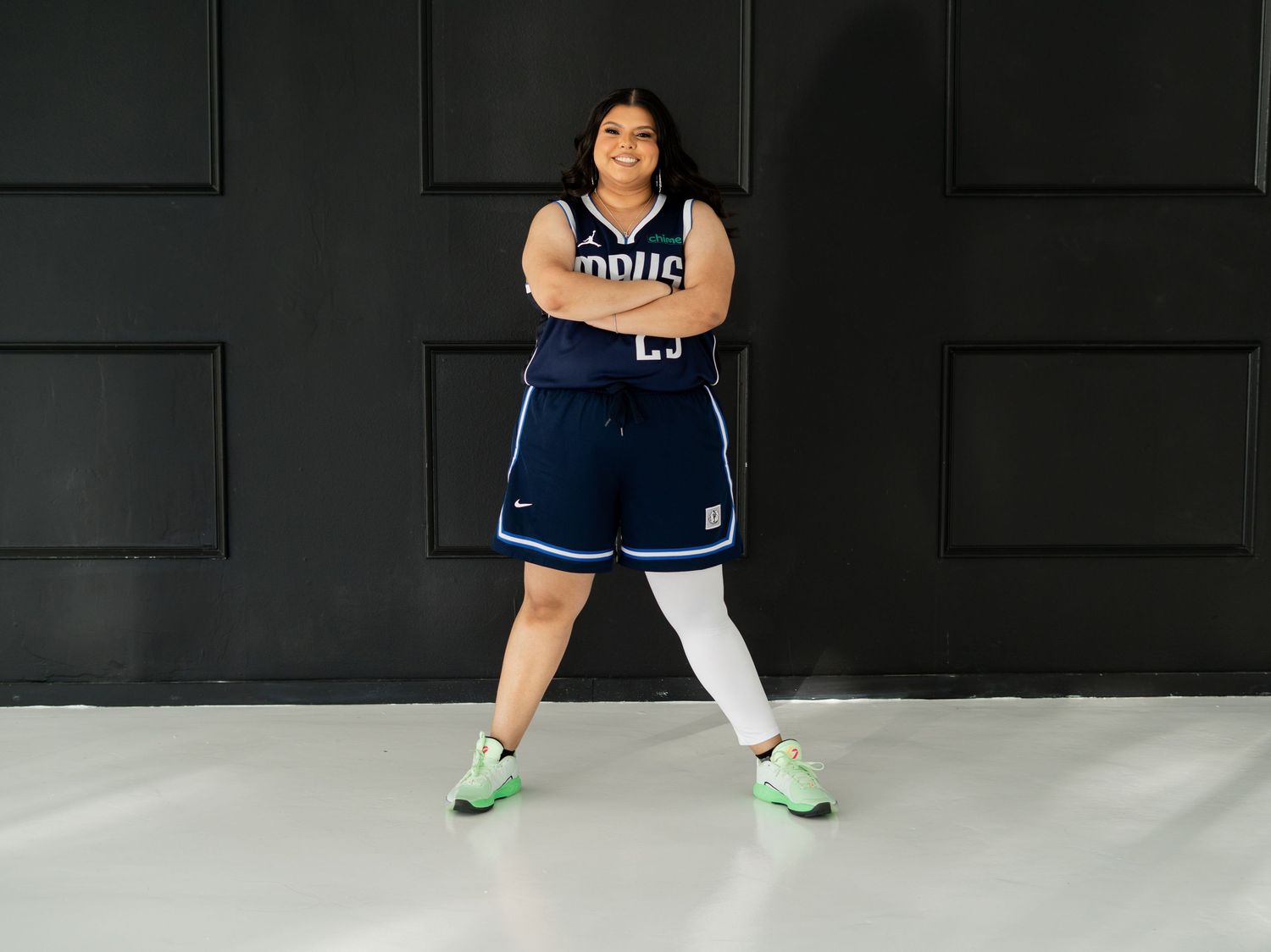 Woman in Mavericks basketball uniform standing confidently in studio with green sneakers.