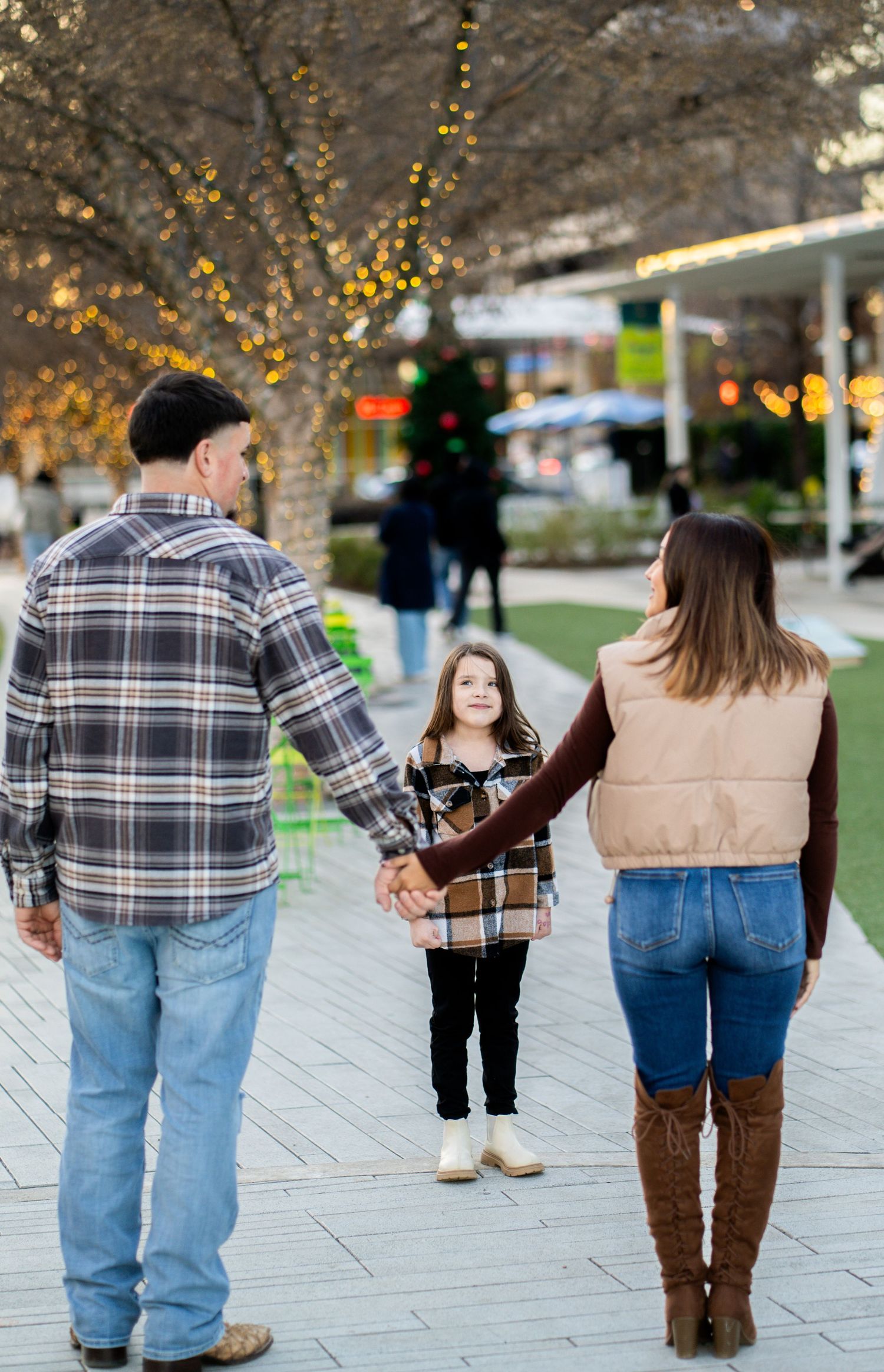 A family of three holds hands while walking along a festive outdoor plaza with holiday lights.