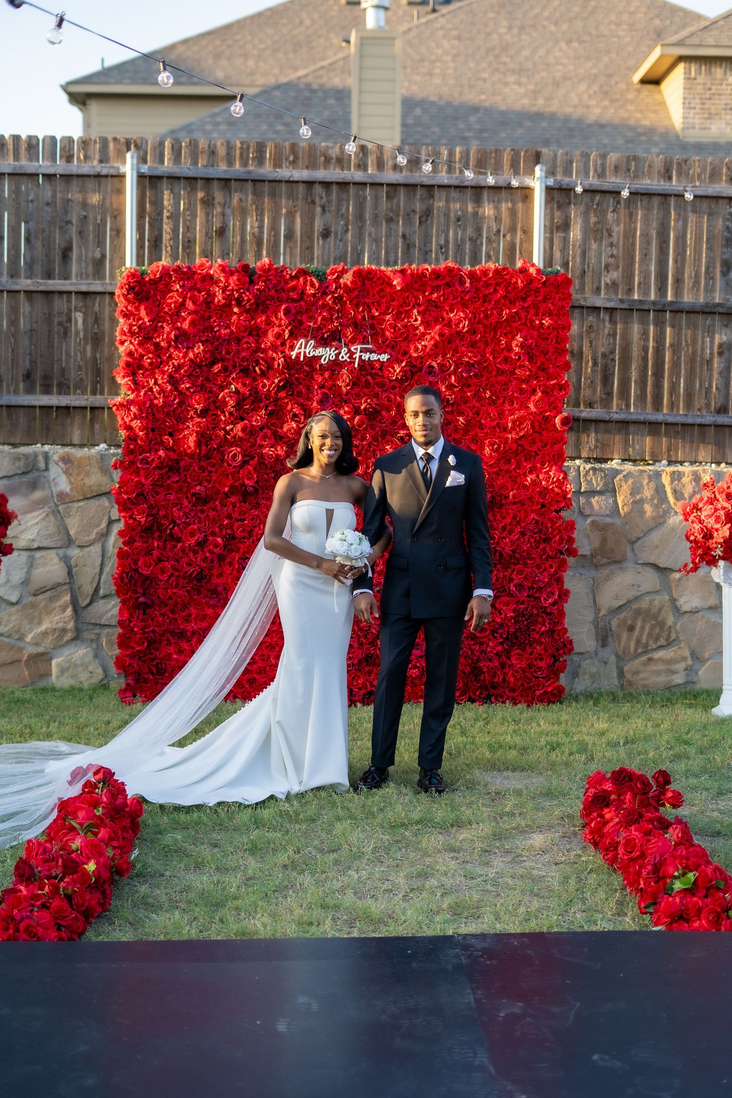 Bride in white gown and groom in black suit pose before red rose wall and floral aisle.