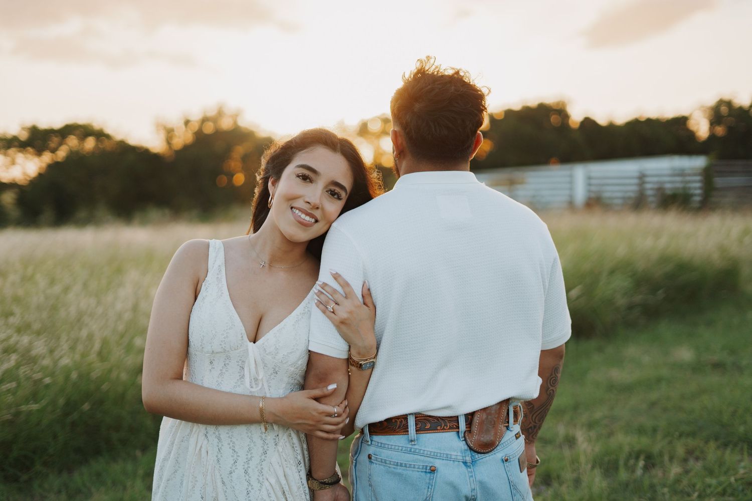 A couple poses at golden hour in a field, the woman in a white dress smiling over her shoulder.