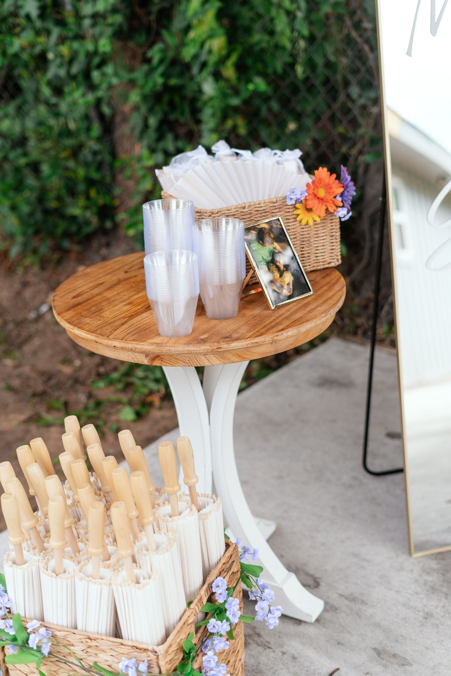 Wooden round table with clear plastic cups, wicker basket with flowers, and white parasols at an outdoor wedding.