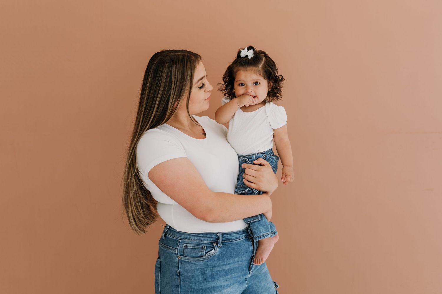 A mother in white top and jeans holds her toddler daughter against a warm terracotta background.