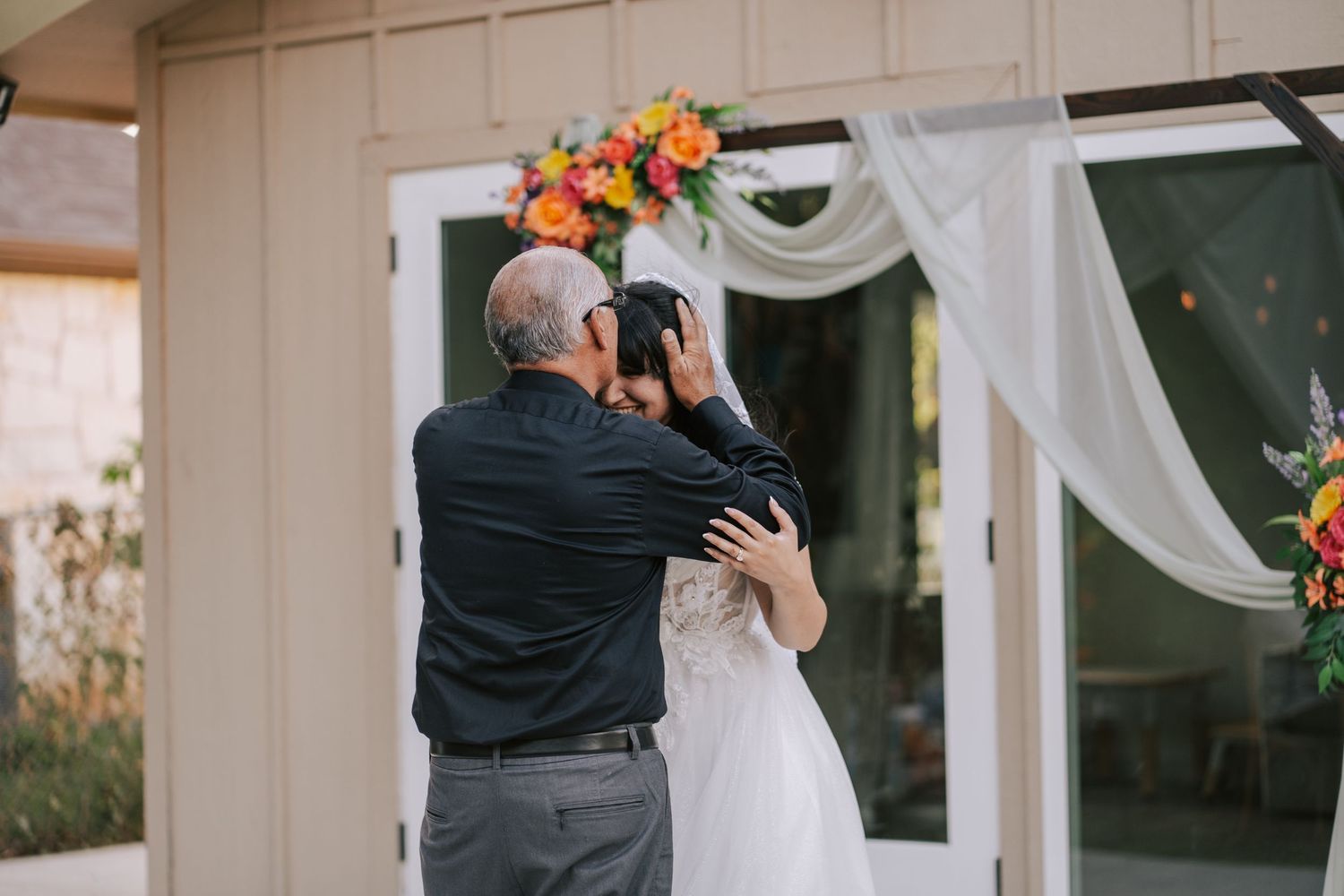 Bride and older man share emotional hug under floral arch at outdoor wedding.
