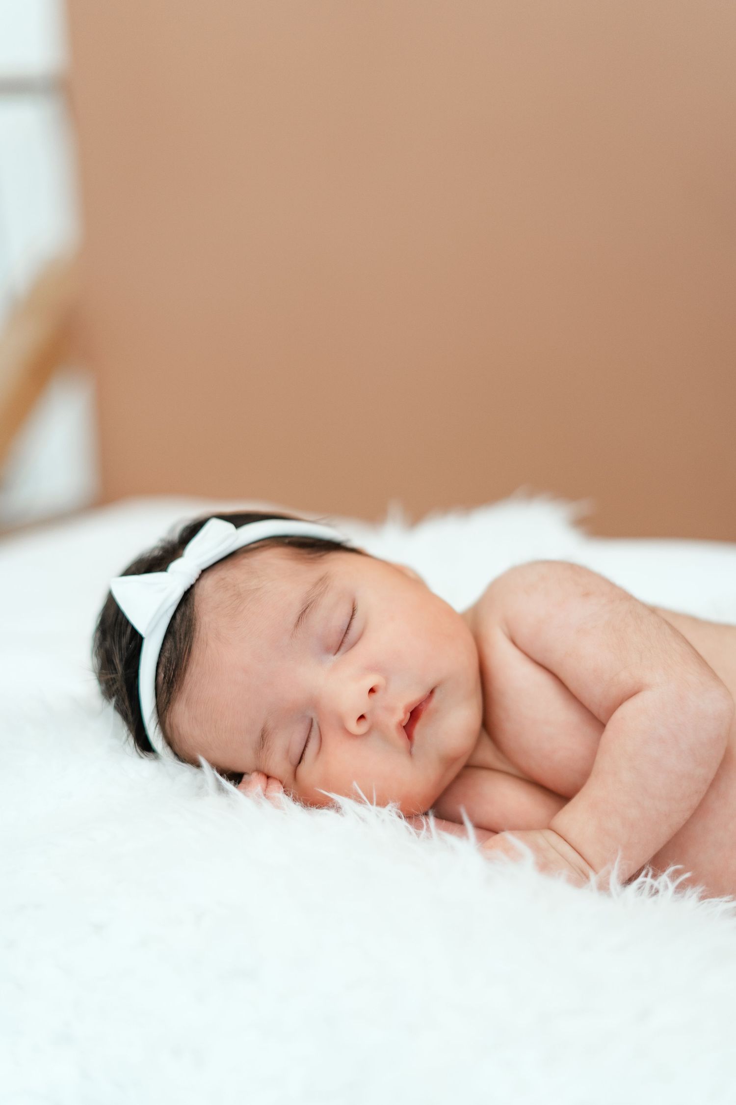 Peaceful newborn baby girl sleeping on white fluffy blanket wearing a delicate white bow headband.