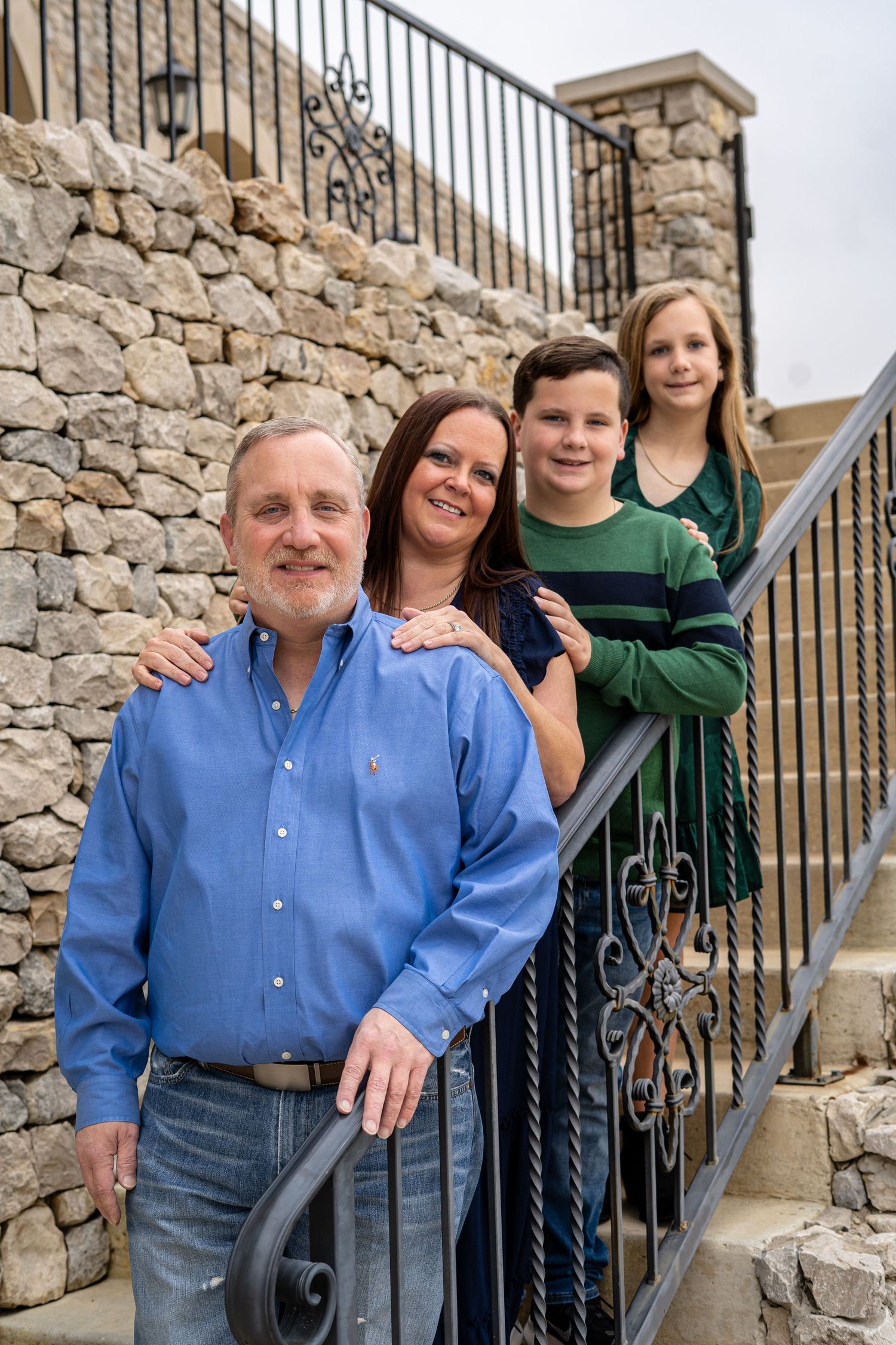 A family of four poses on stone steps with an ornate iron railing against a rustic stone wall.