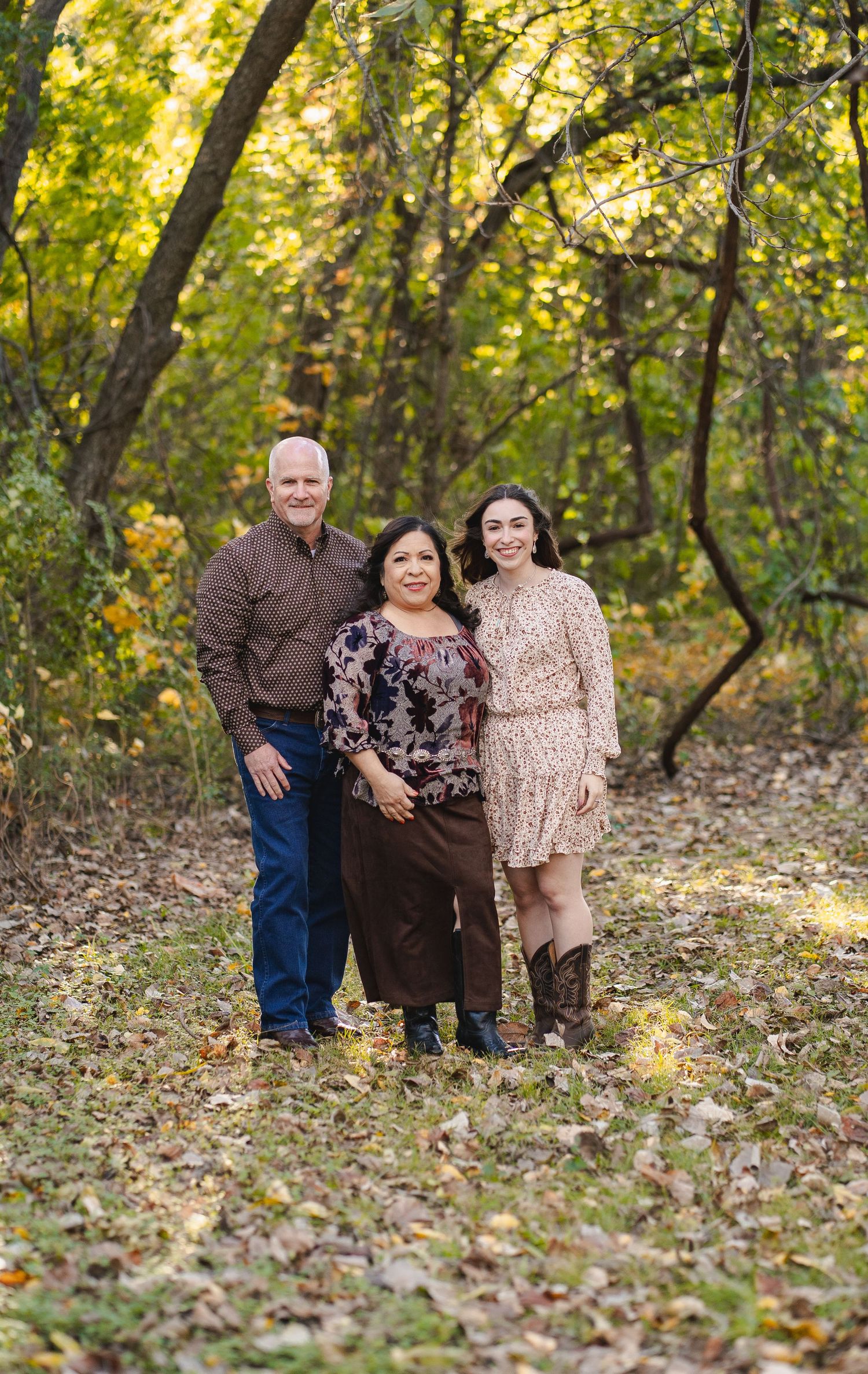 Three family members smiling together in a wooded area with colorful autumn foliage.