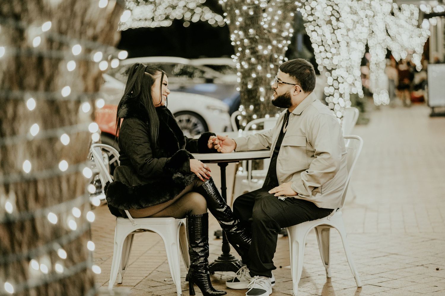 A couple sits at an outdoor cafe table holding hands surrounded by sparkling holiday lights.