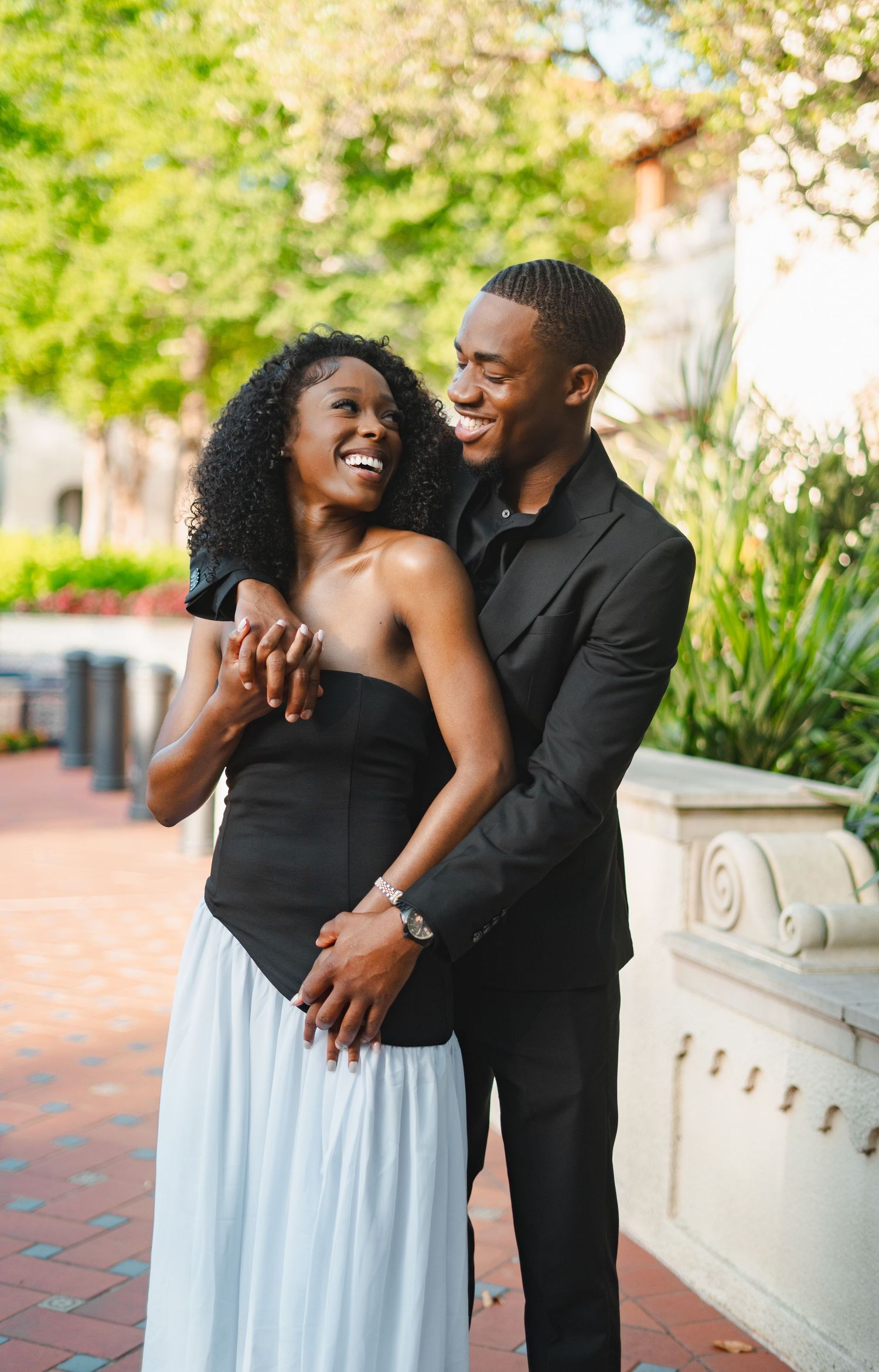 Elegant Black couple laughing and embracing outdoors, wearing black formal attire in a garden setting.