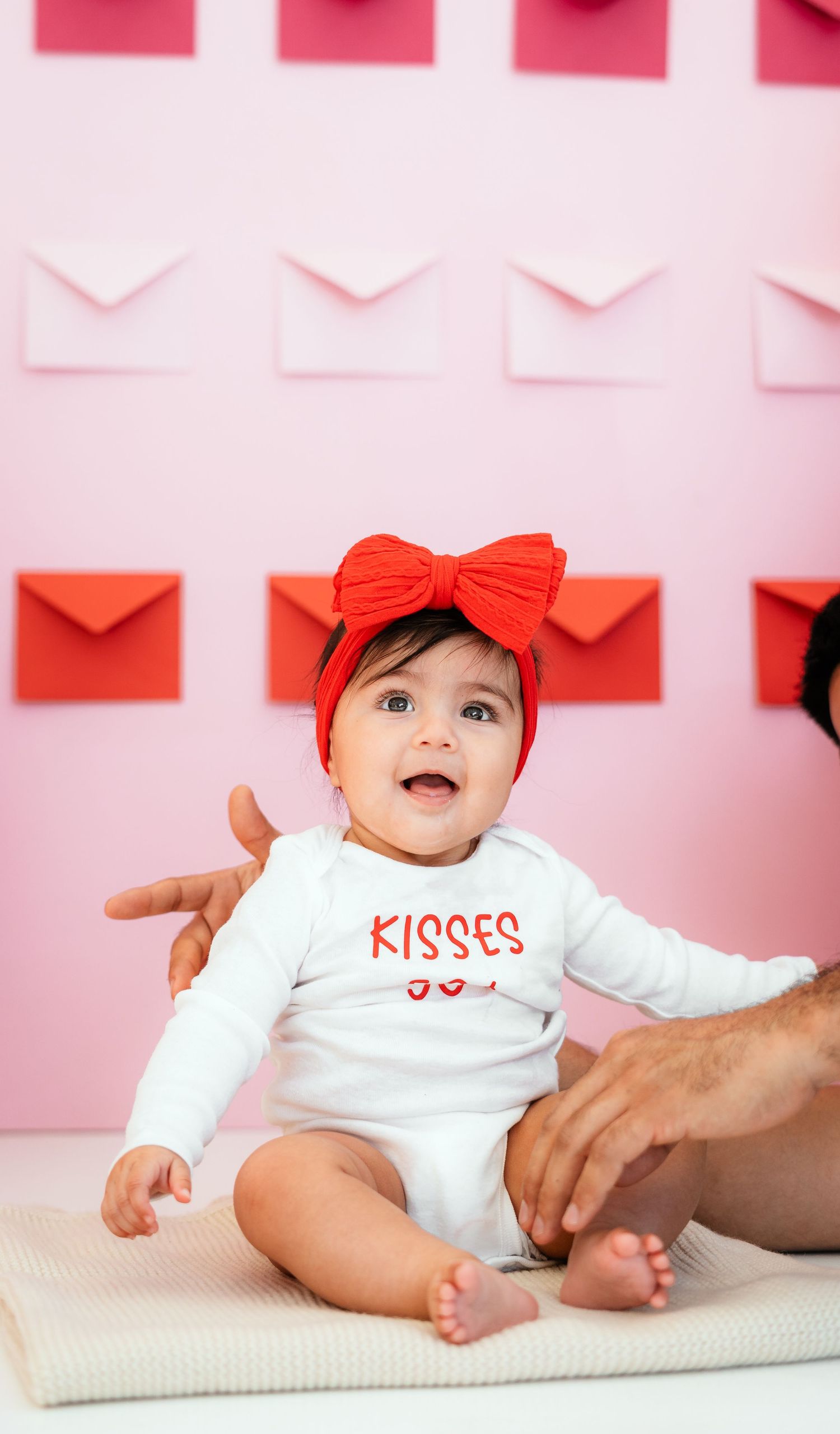 Baby in white Kisses onesie with red bow headband sits before pink envelope wall backdrop.