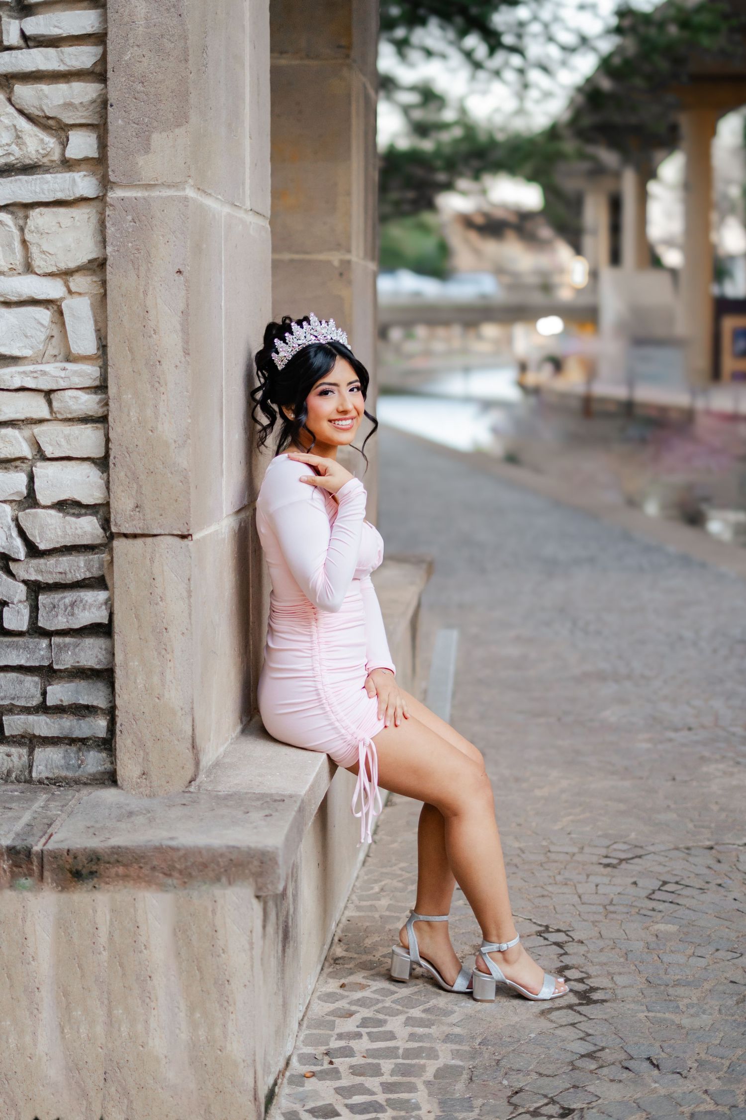 Young woman in pink mini dress and tiara sits gracefully on stone ledge, posing for quinceañera photos outdoors.