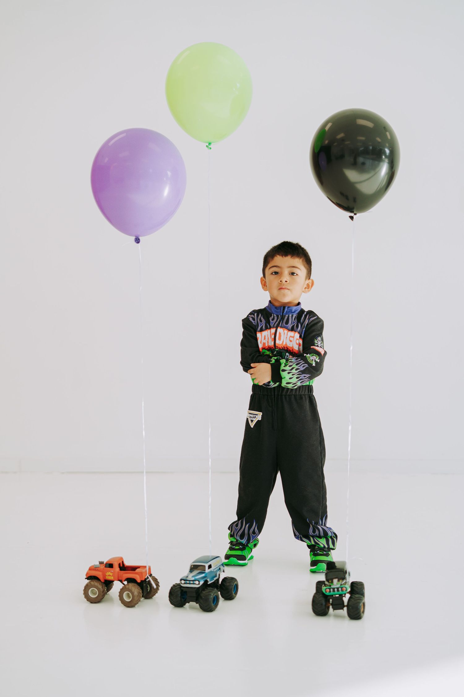 Boy in Monster Jam costume stands with colorful balloons and monster truck toys on white studio floor.