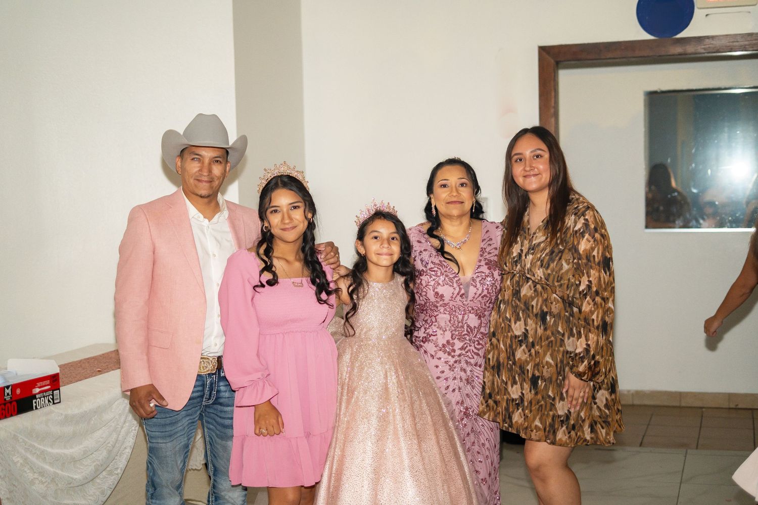 Quinceañera girl in pink gown poses with family including father in pink cowboy hat at her party.