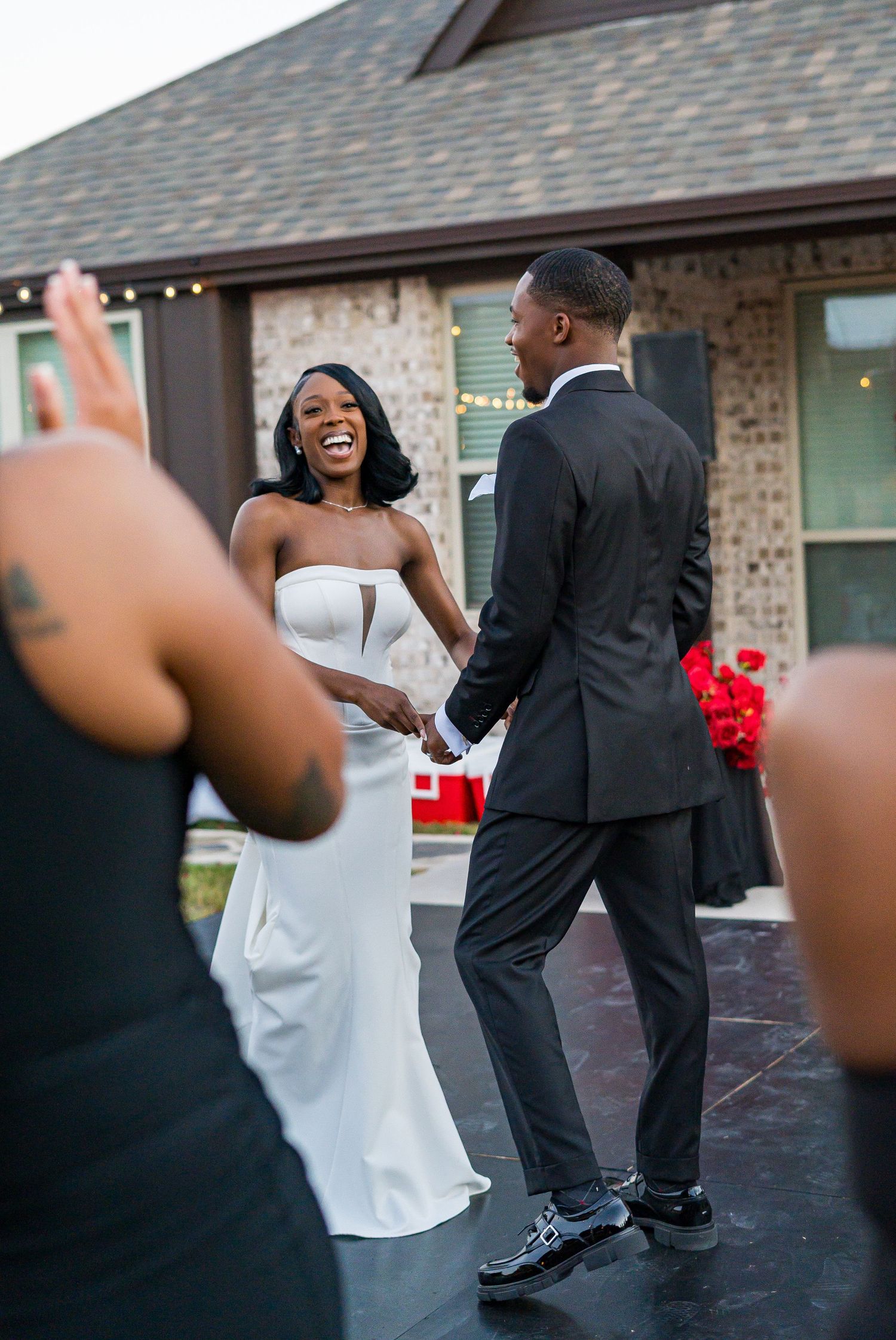 Laughing bride and groom hold hands while dancing on outdoor stage at evening wedding reception.