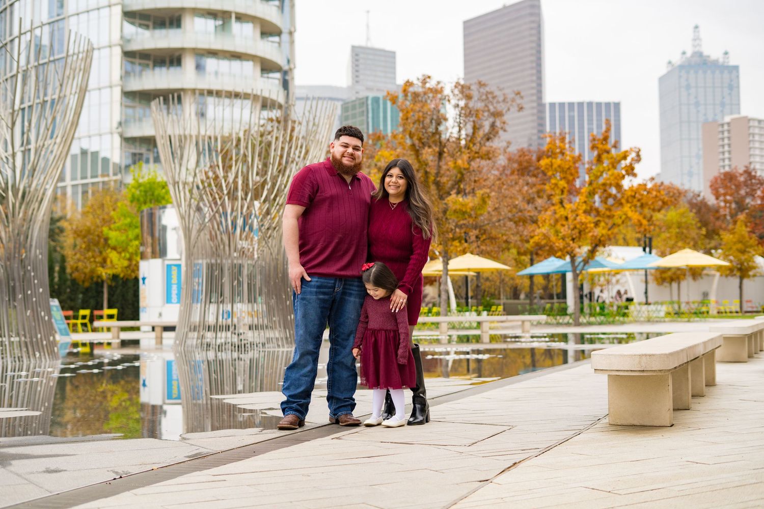 Family of three posing by a reflective pool with a city skyline backdrop in autumn.