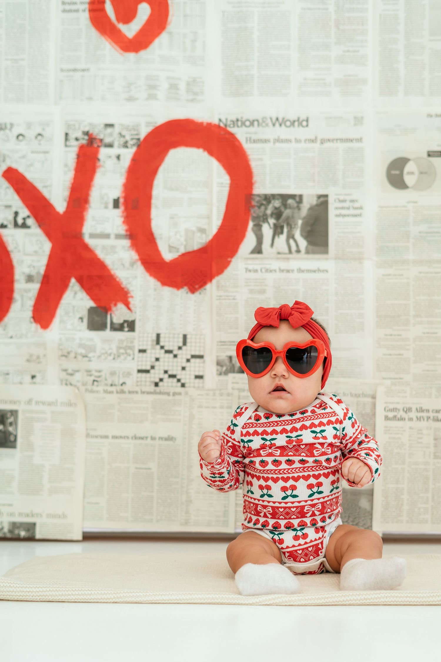 Baby in heart sunglasses and red bow sits before Valentine's XOXO backdrop.