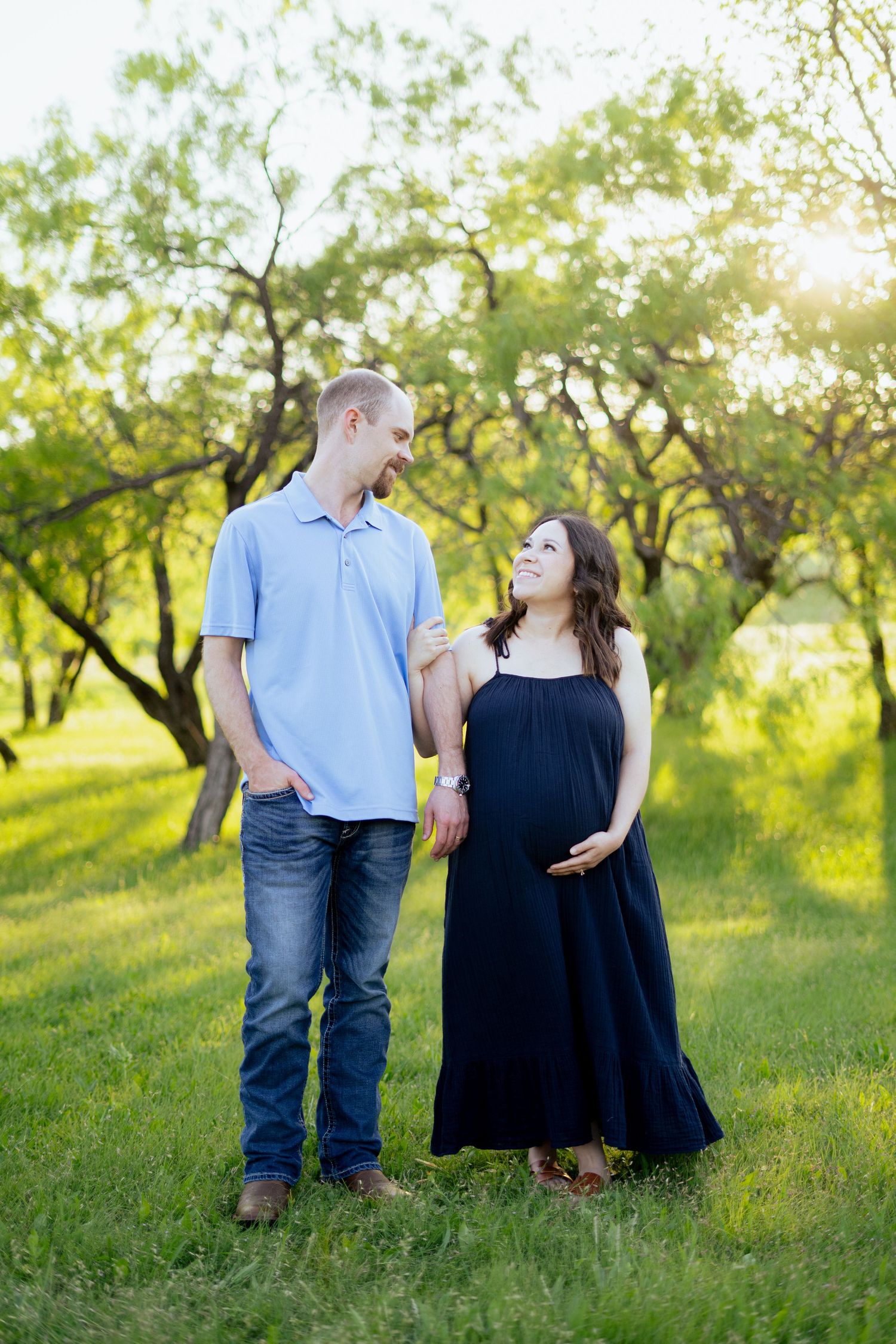 Pregnant couple holding hands and smiling at each other in a sunny green park.