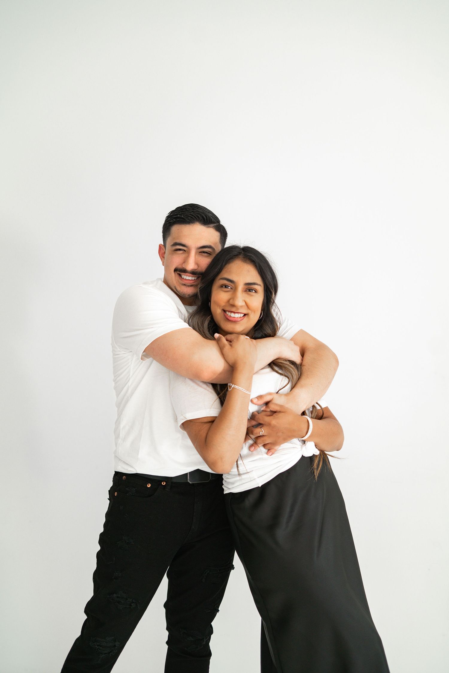 Man in white tee hugging woman from behind, both smiling, against a white background.