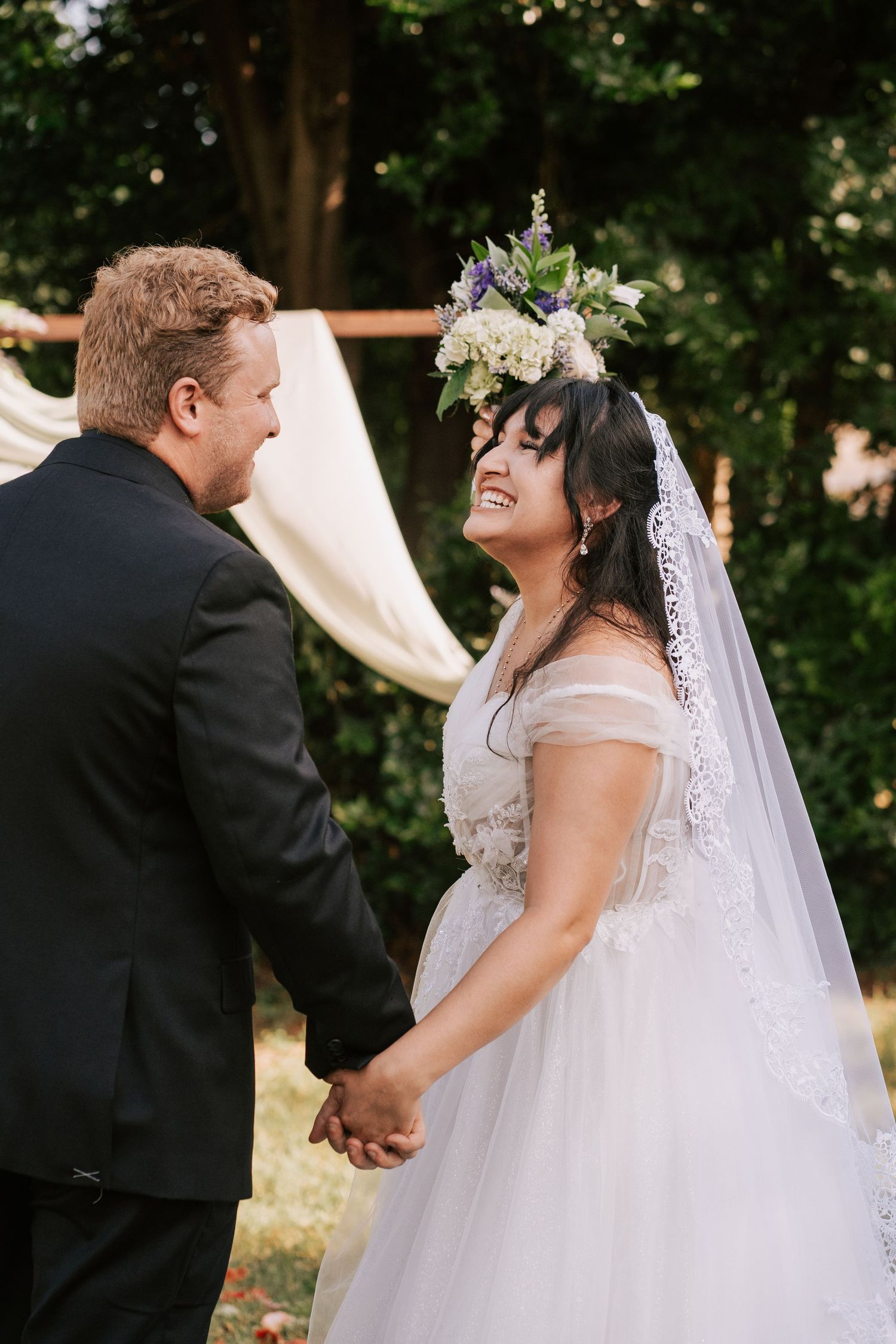 Bride in floral crown and veil laughs joyfully holding hands with groom in black suit at outdoor wedding ceremony.