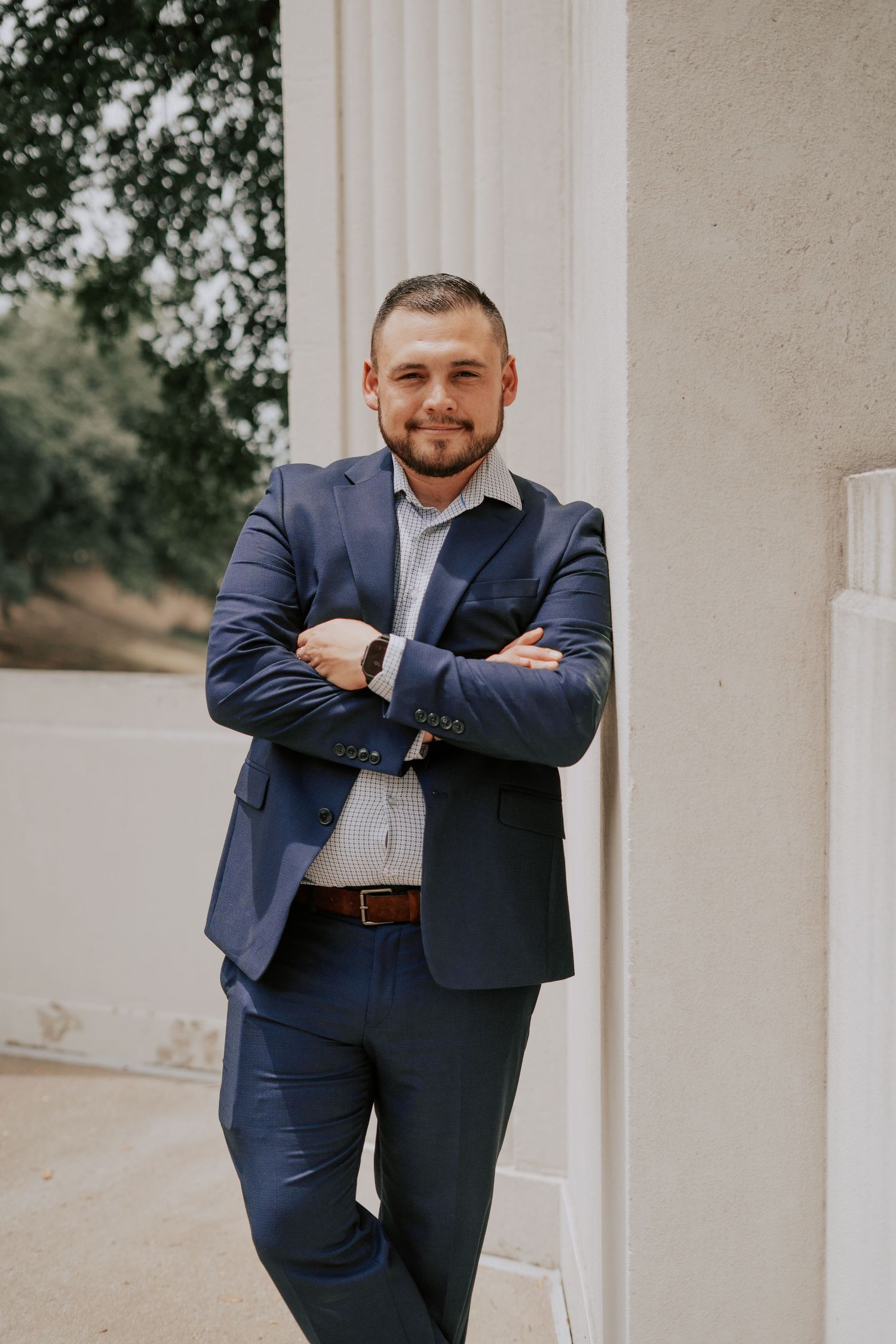 Professional man in navy suit standing with arms crossed leaning against white pillar outdoors.