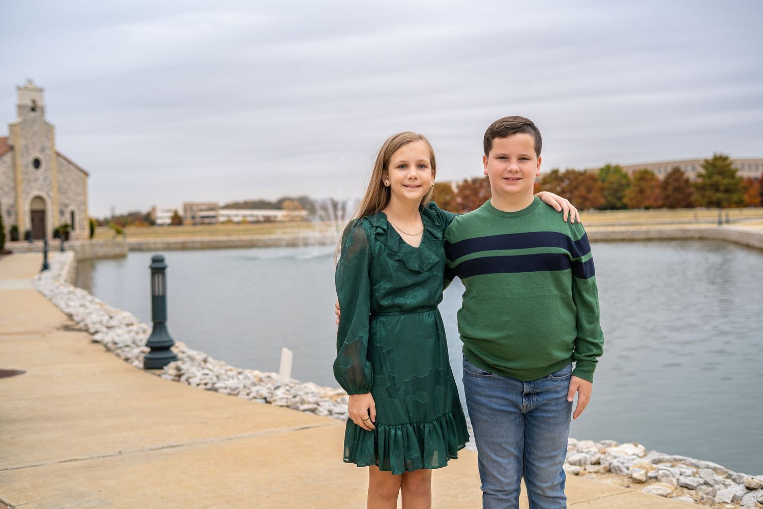 A young girl in green dress and boy in striped sweater posing together by a lakeside stone path.