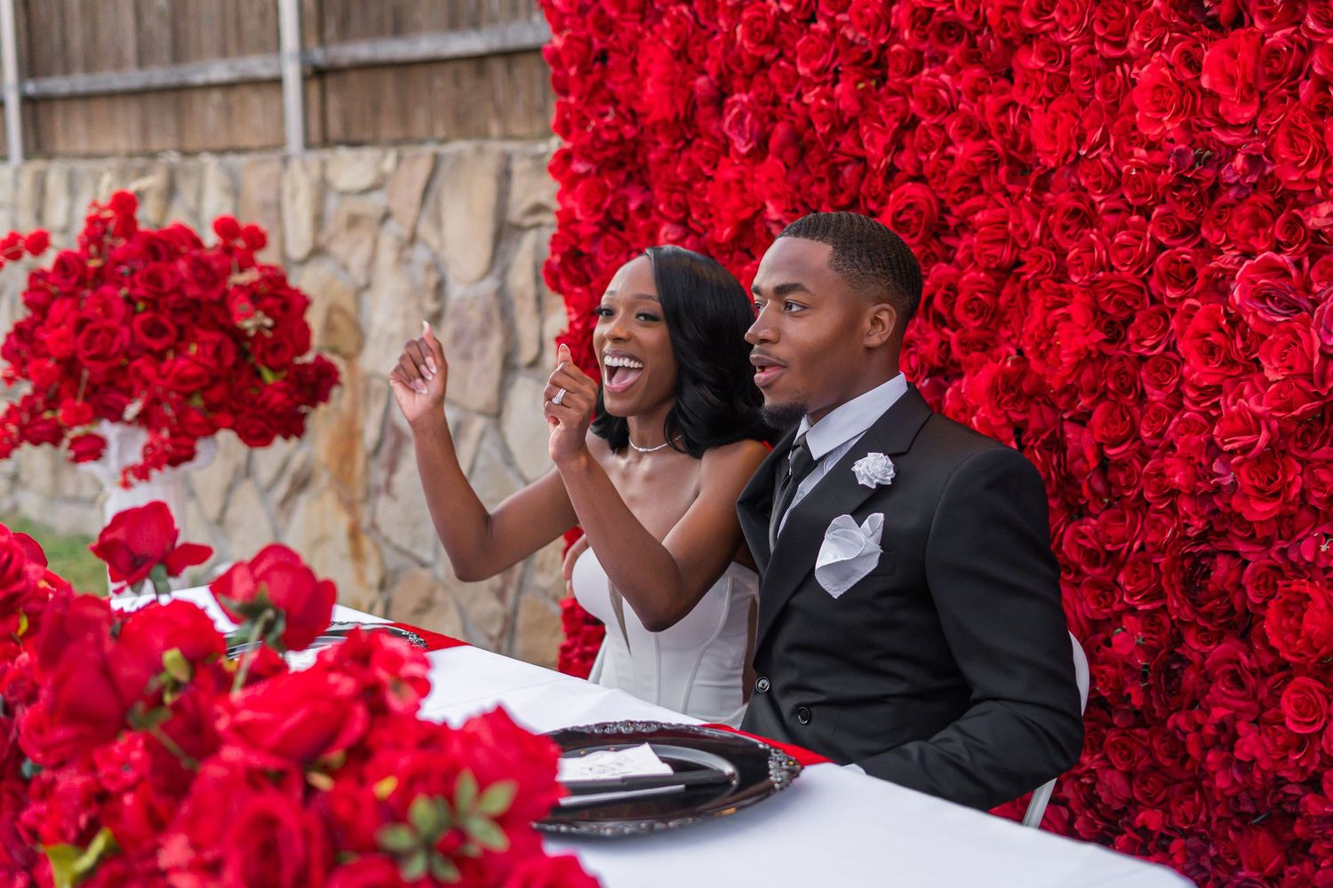 Bride and groom share a joyful moment at their sweetheart table adorned with red floral arrangements.
