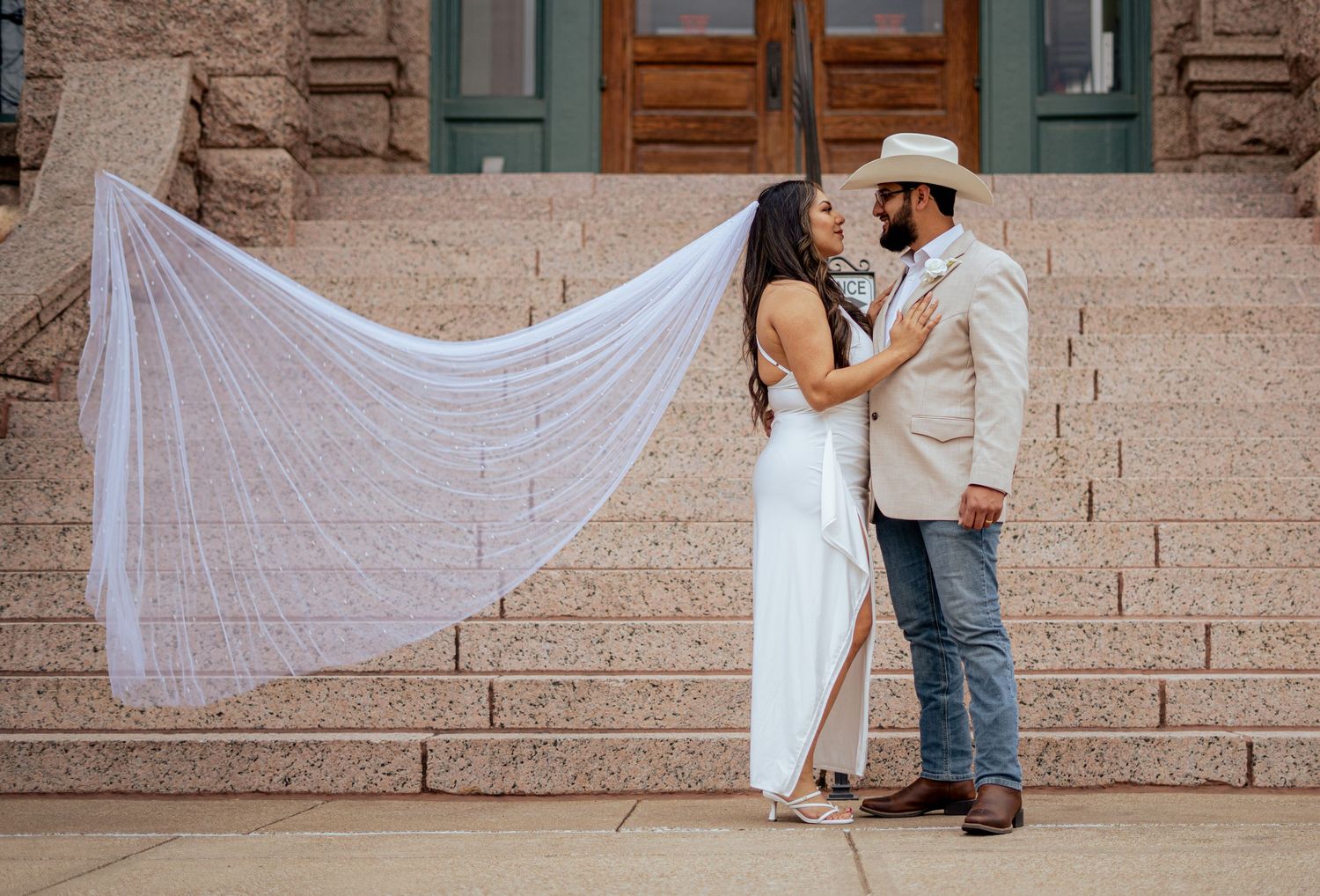 Bride's long veil flows dramatically in the wind as couple shares romantic moment on stone steps.