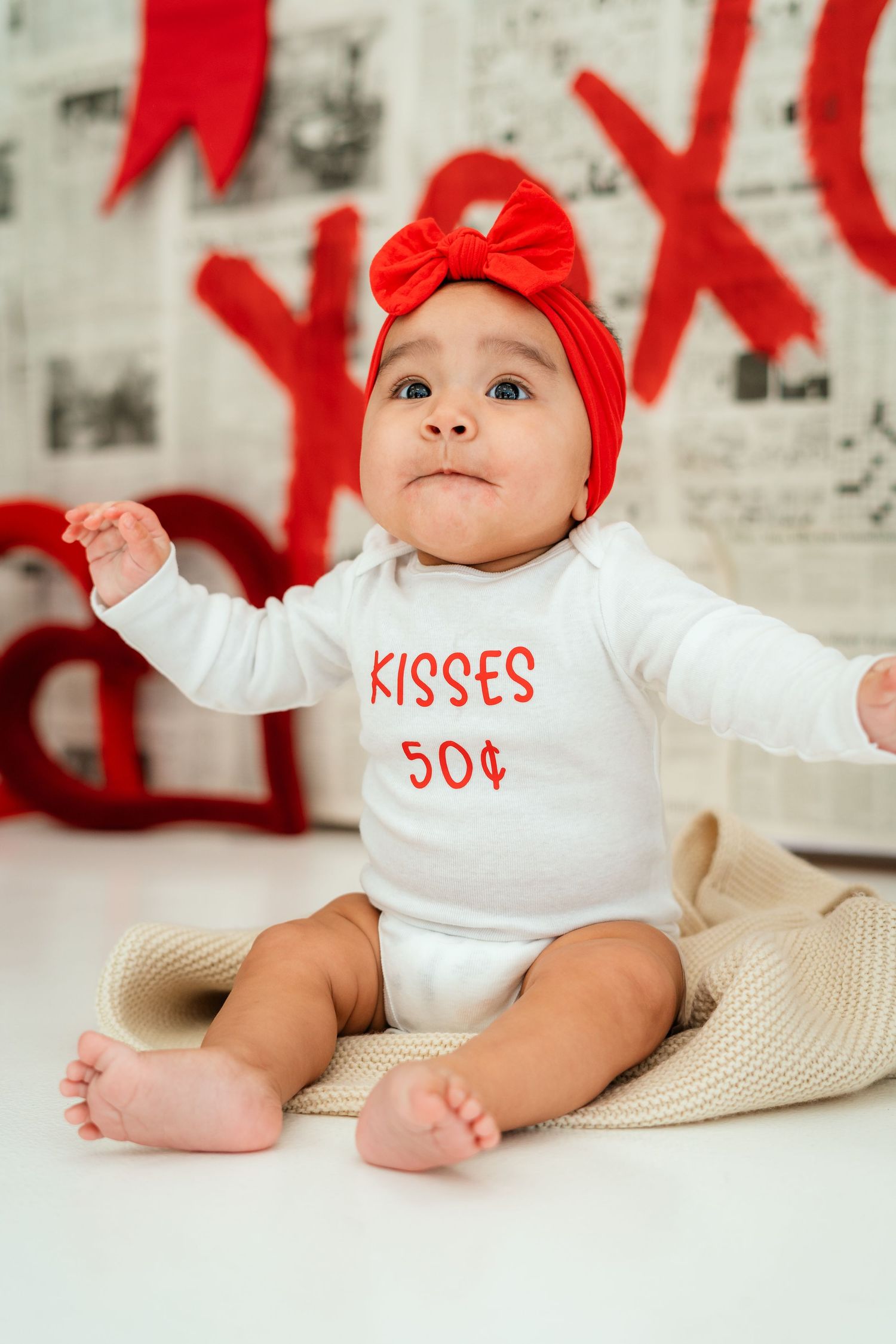 Baby in white 'Kisses 50 cents' onesie with red bow before XOXO backdrop.