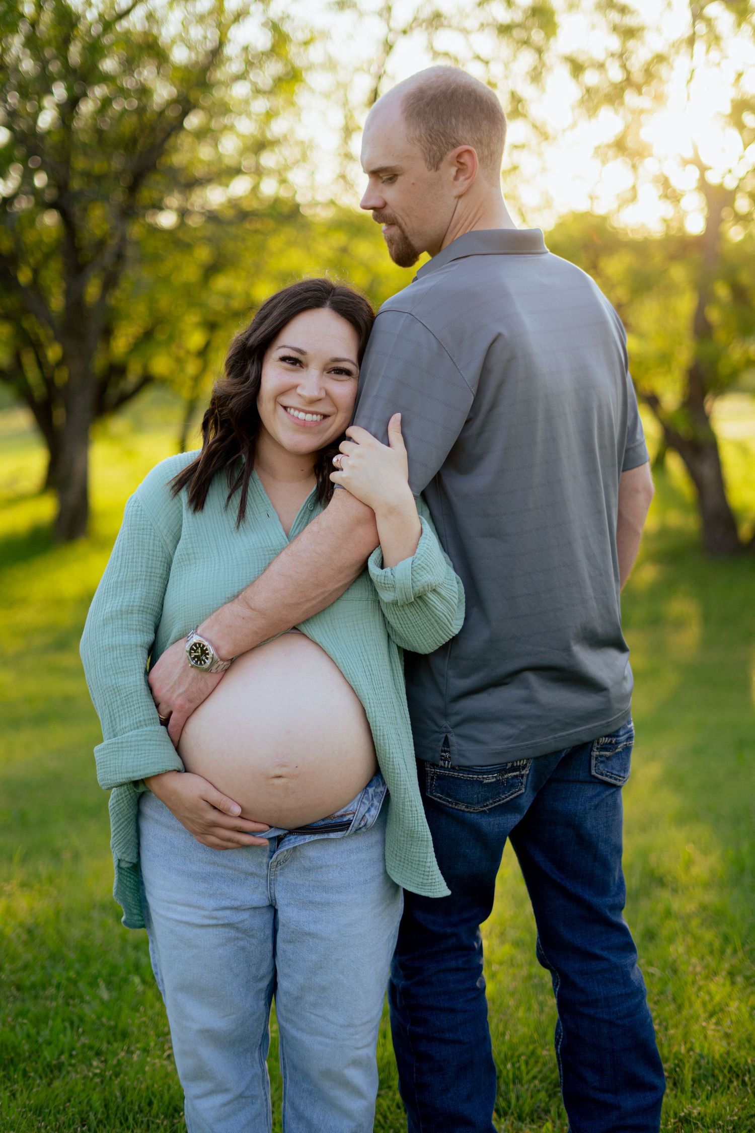 Smiling pregnant woman in mint green shirt embraces partner outdoors in sunny green park during maternity photo session.