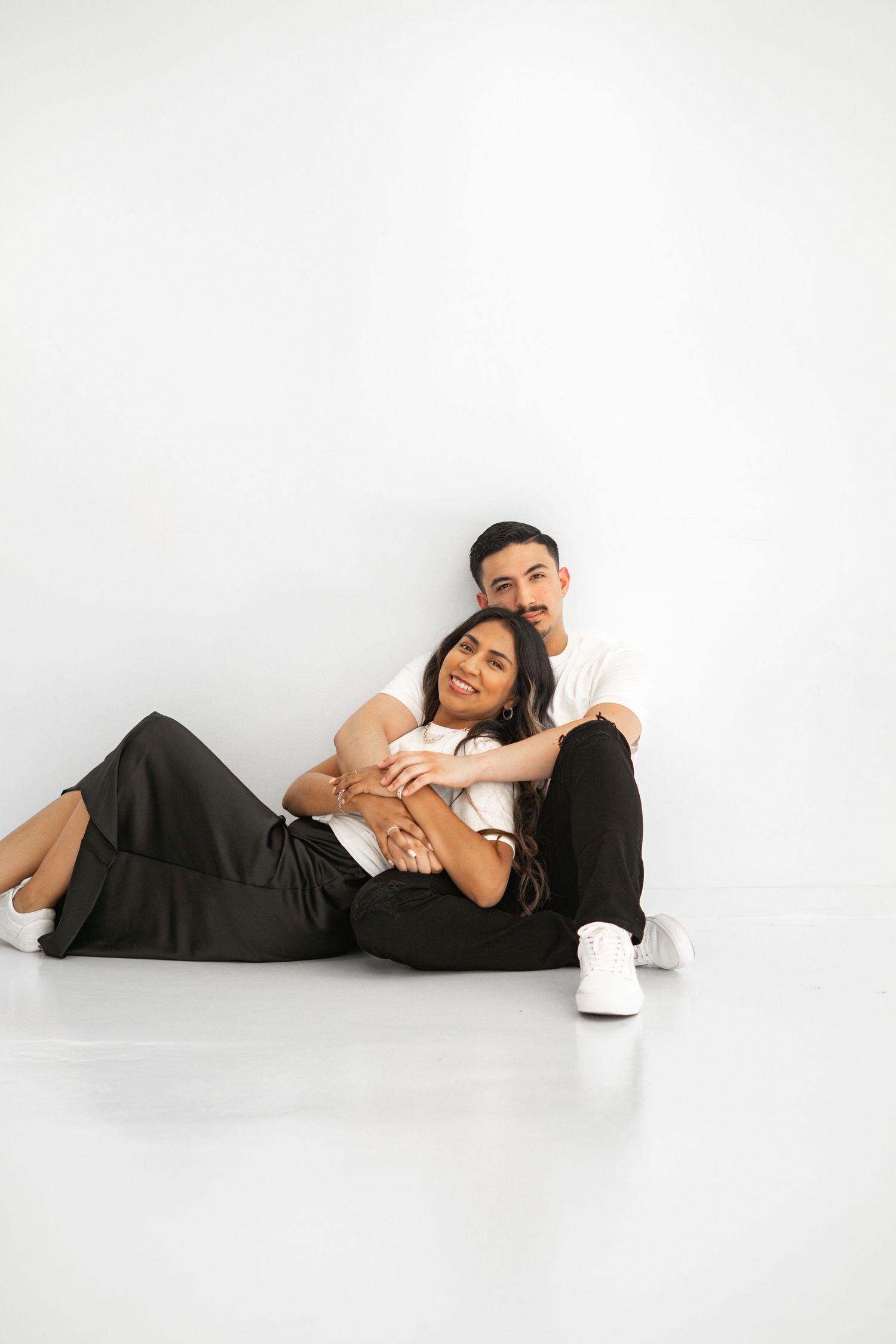 Couple sitting on floor, woman reclining on mans lap, smiling at camera on white background.