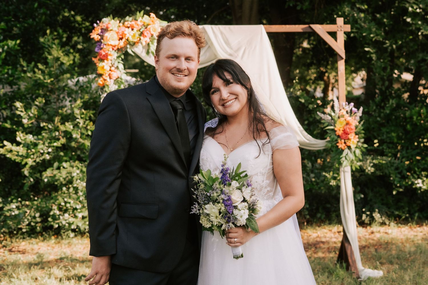 Smiling bride and groom pose before floral arch at outdoor wedding ceremony.