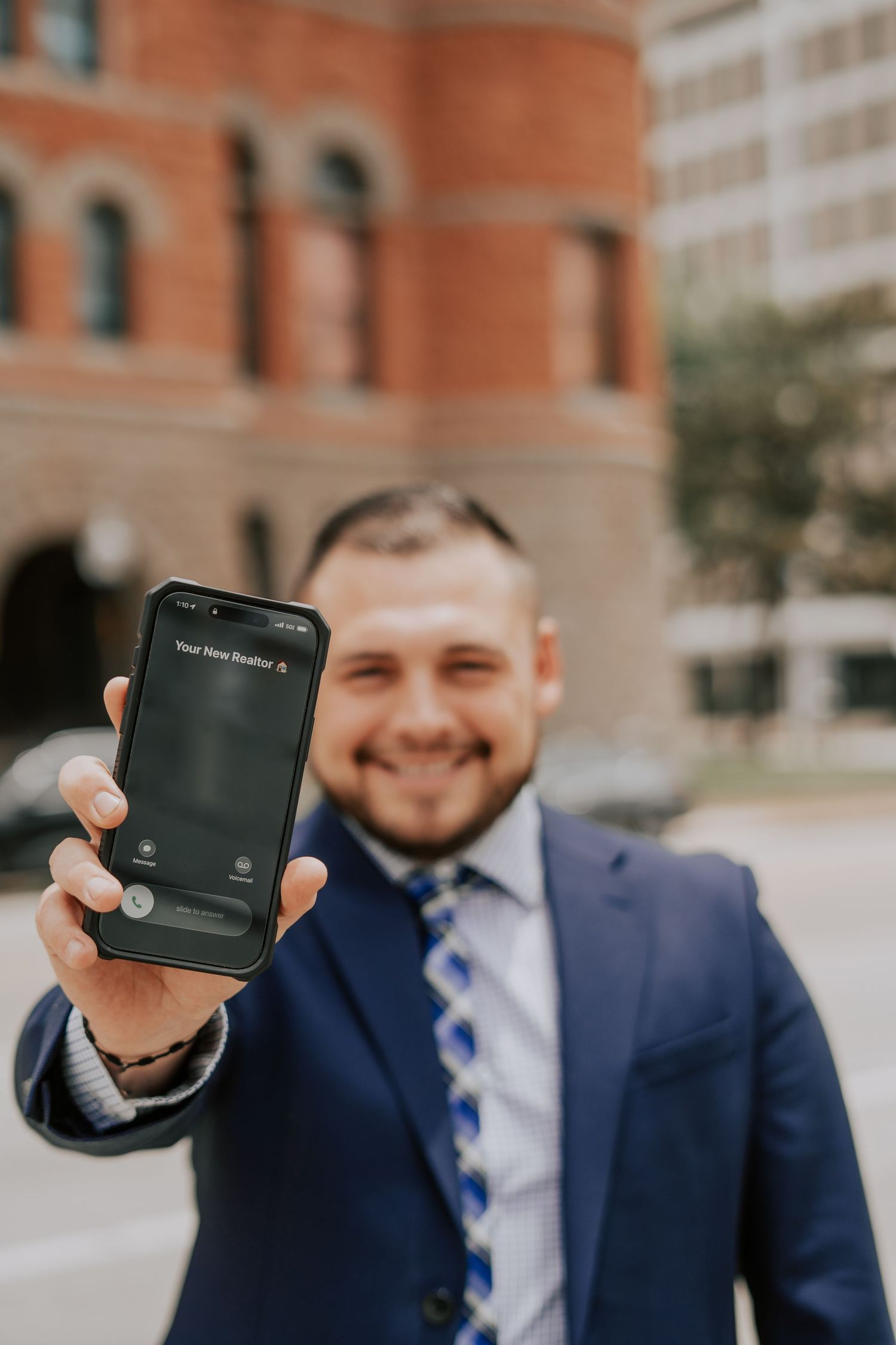 Smiling businessman in navy suit holding smartphone screen toward camera on urban city street.
