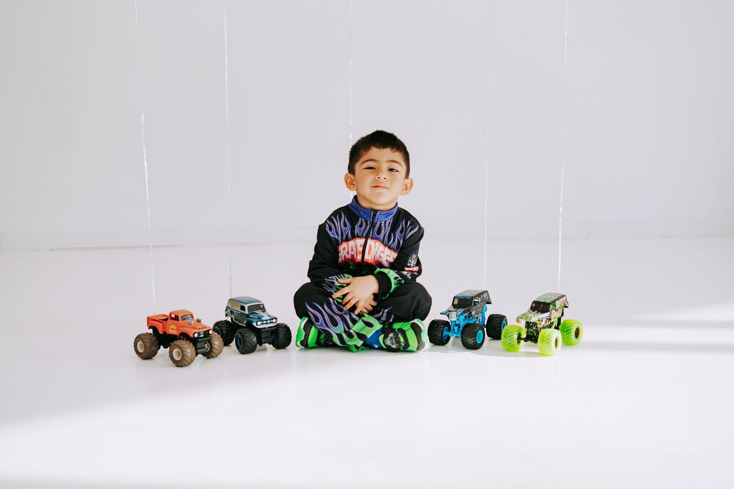 Smiling boy in Monster Jam outfit sits on floor surrounded by collection of colorful monster truck toys.