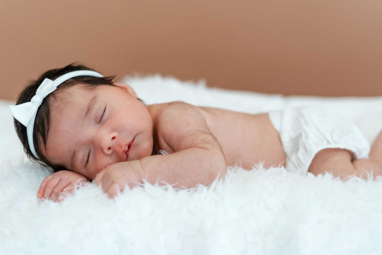 Newborn baby girl in white diaper sleeping on soft white fur blanket with white bow headband.