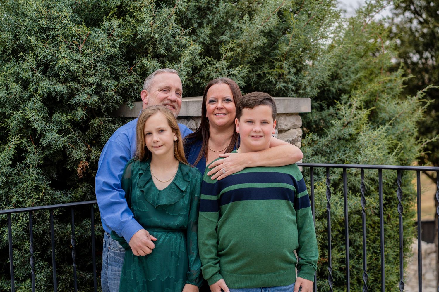 A family of four smiles warmly together in front of lush green bushes on a wooden deck.