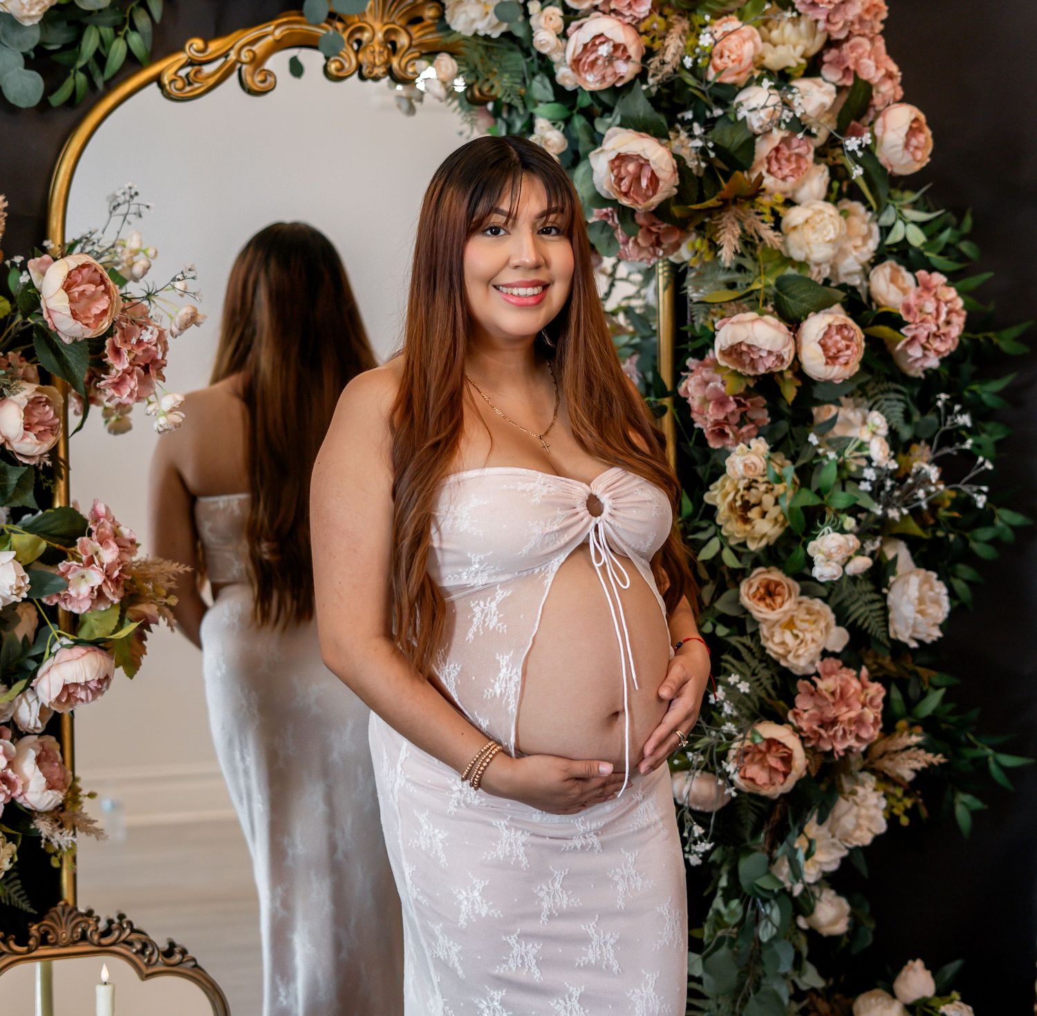 Smiling pregnant woman in white lace dress holding belly, standing before a floral arch with mirror reflection.
