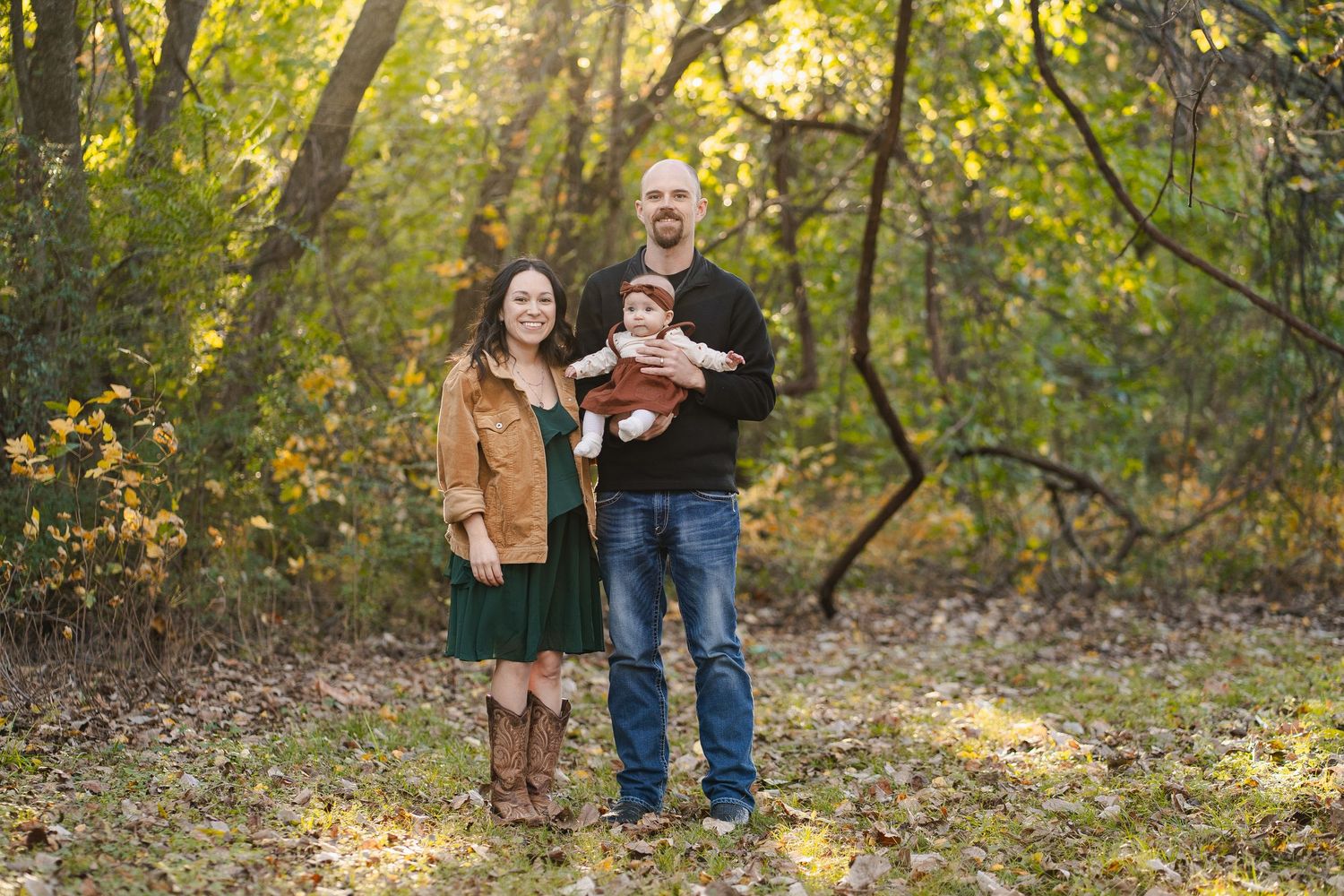 Couple standing together in an autumn forest holding their newborn baby in warm golden light.